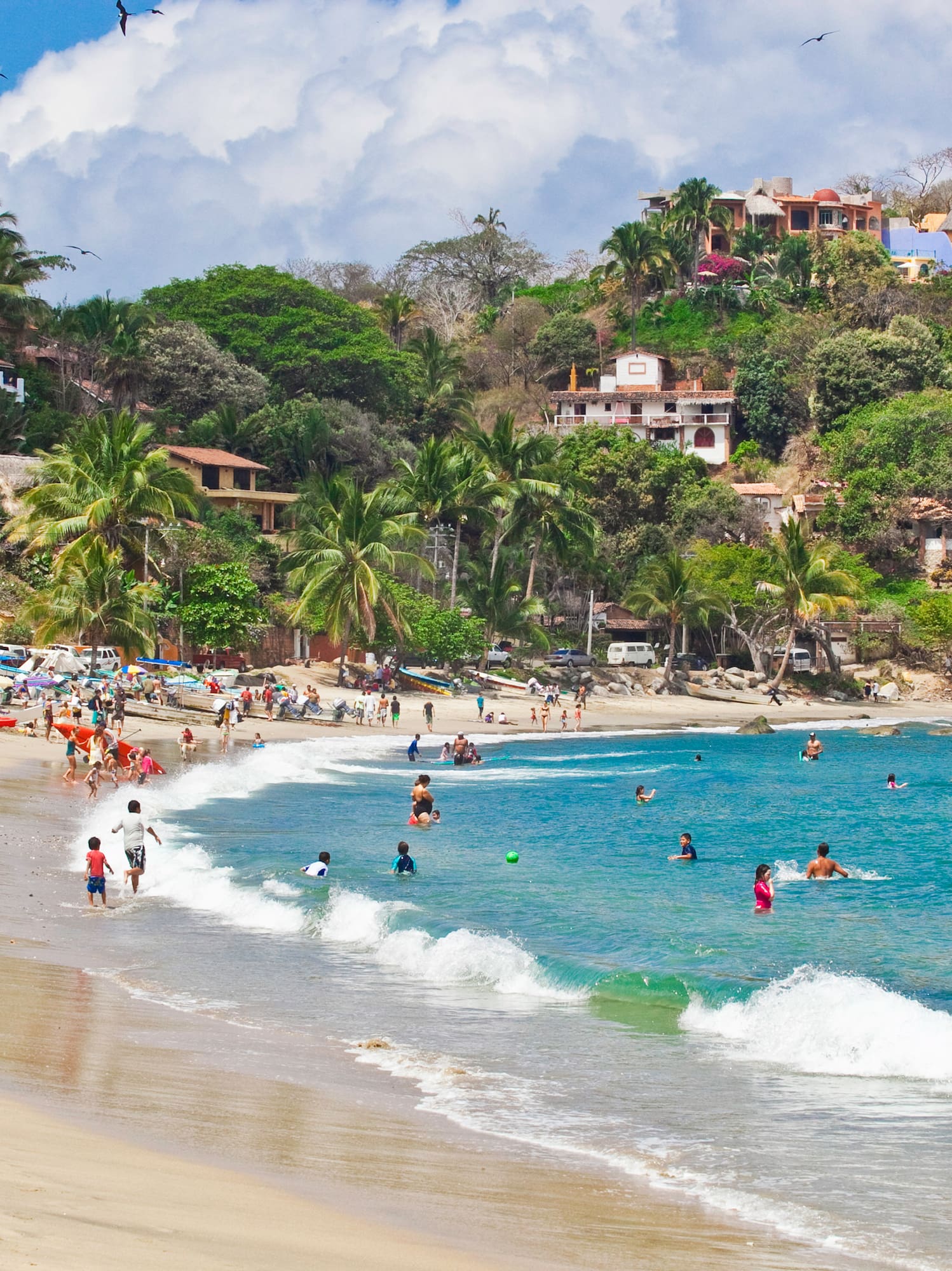 Tropical beach with people swimming, hillside village, palm trees, and blue ocean.