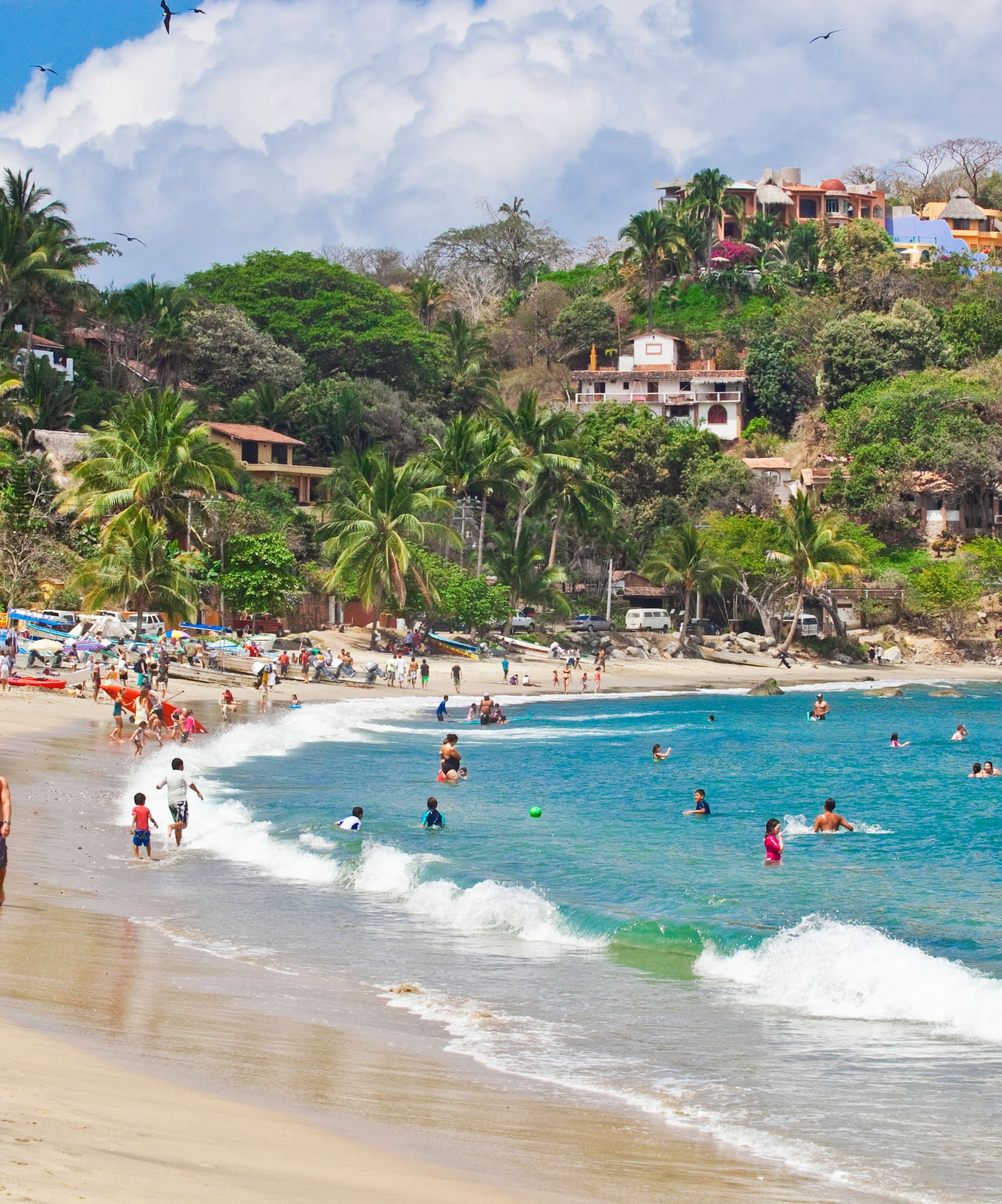 Tropical beach with people swimming, hillside village, palm trees, and blue ocean.