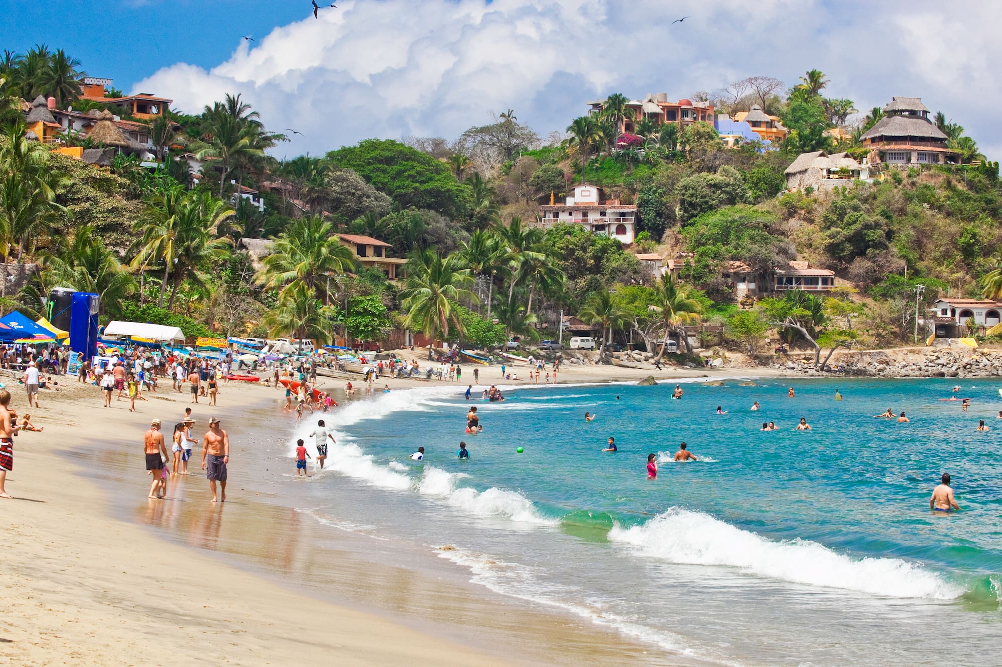 Tropical beach with people swimming, hillside village, palm trees, and blue ocean.