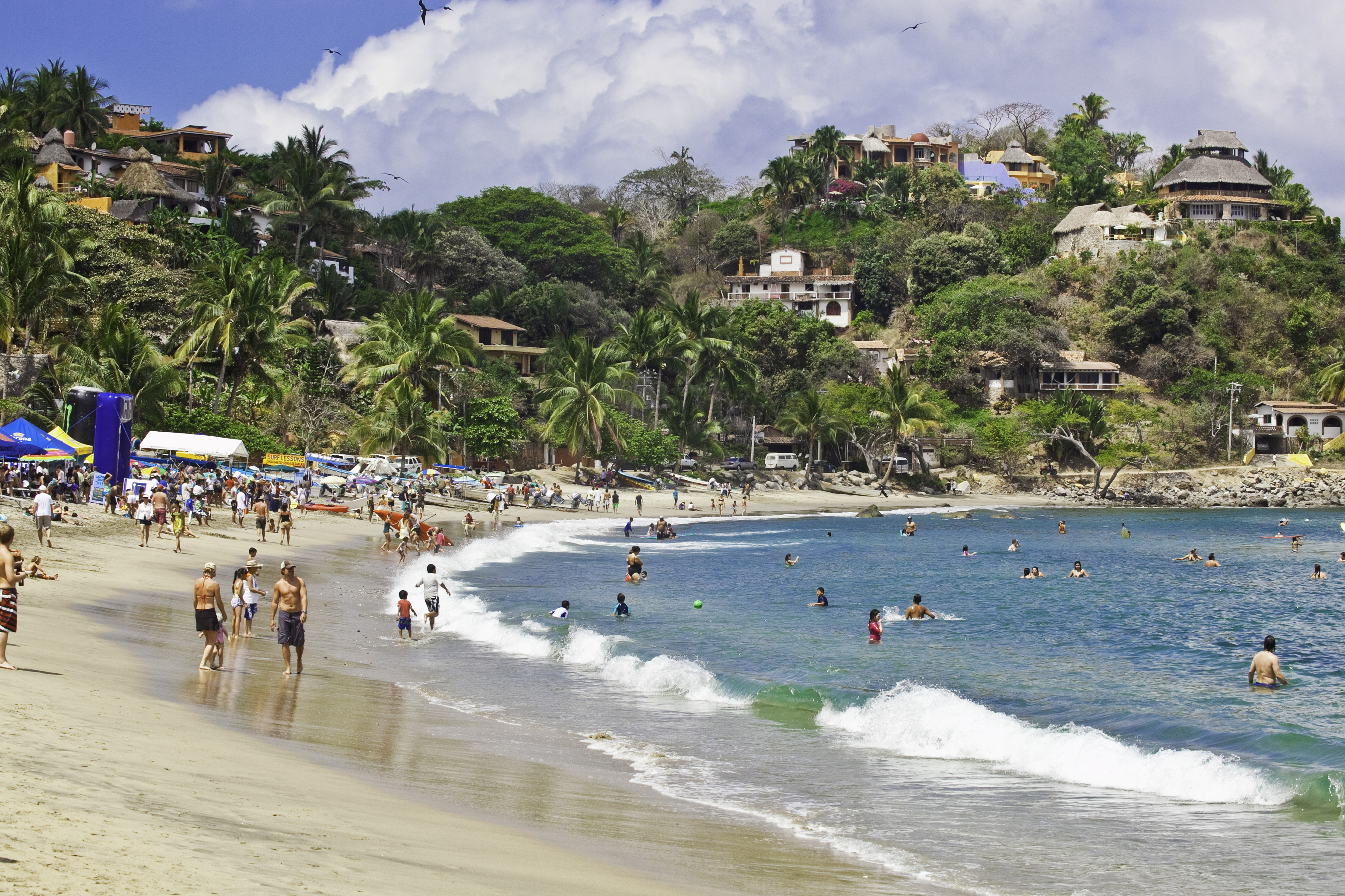 Tropical beach with people swimming, hillside village, palm trees, and blue ocean.