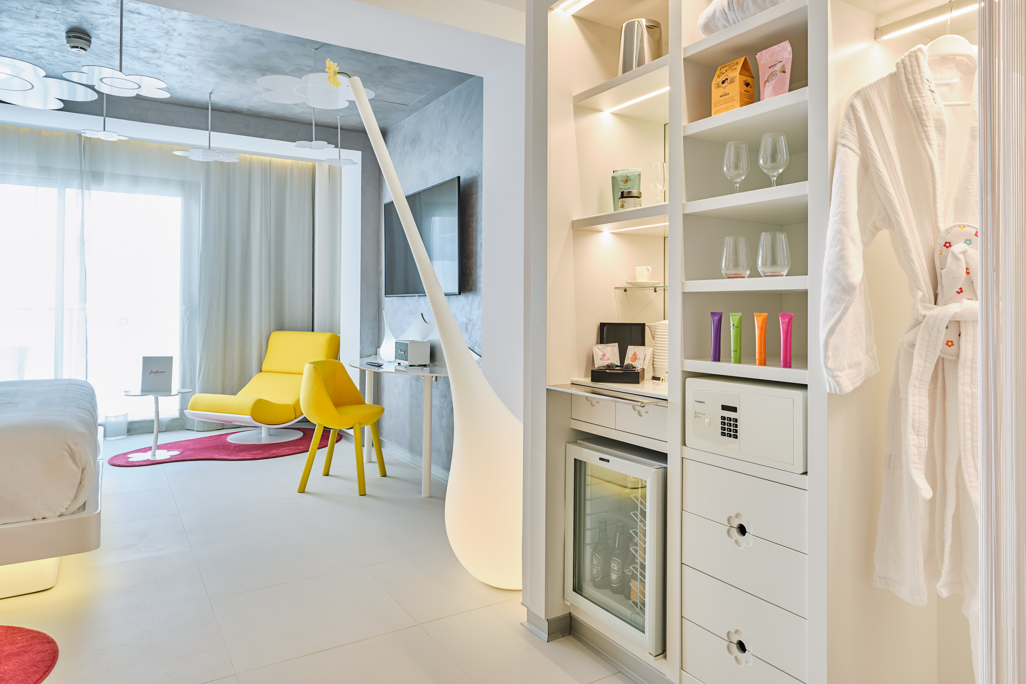 Modern hotel room with yellow chair, white bed, and built-in closet with mini-bar.