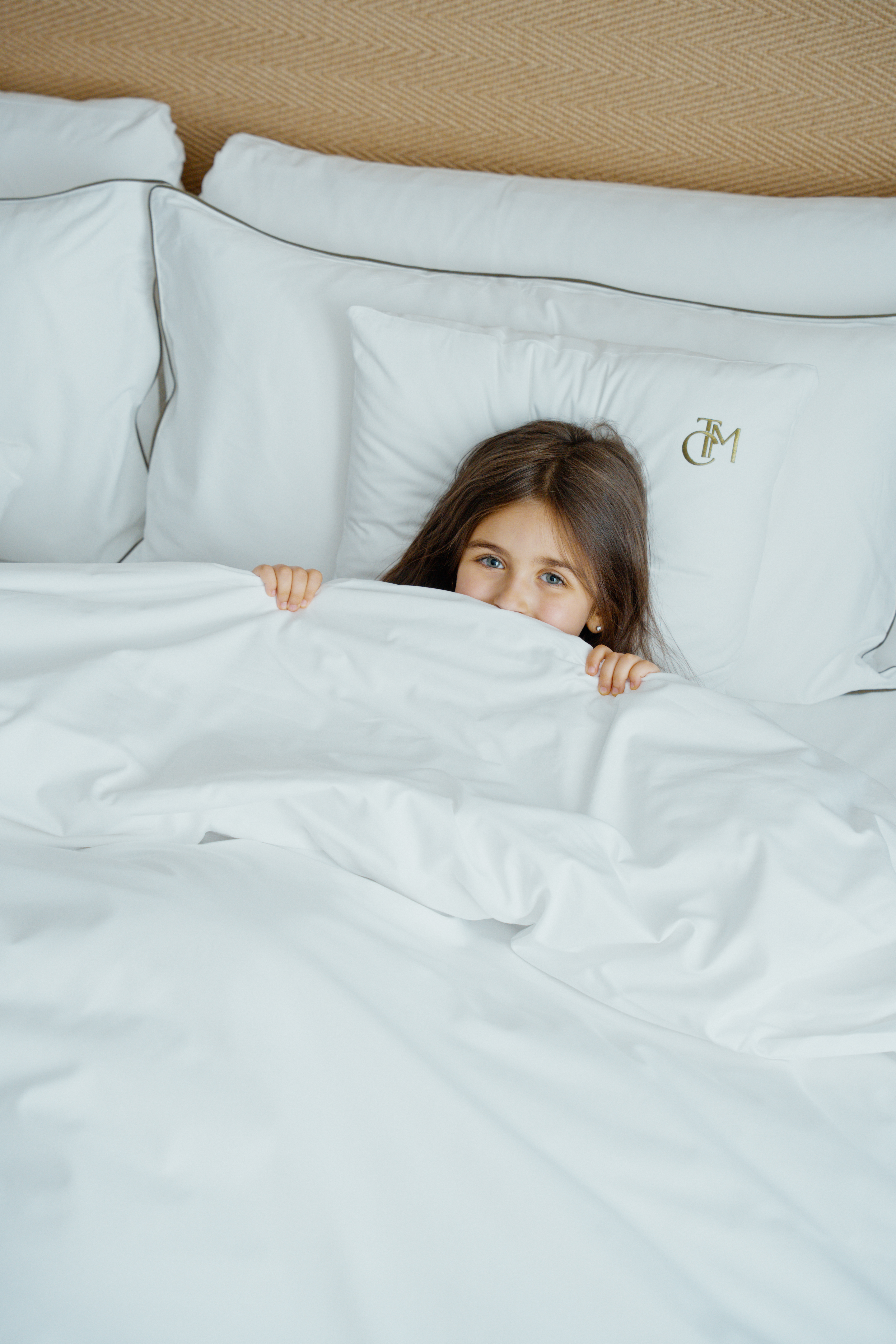 Young girl peeking playfully from under white bed covers with monogrammed pillows.