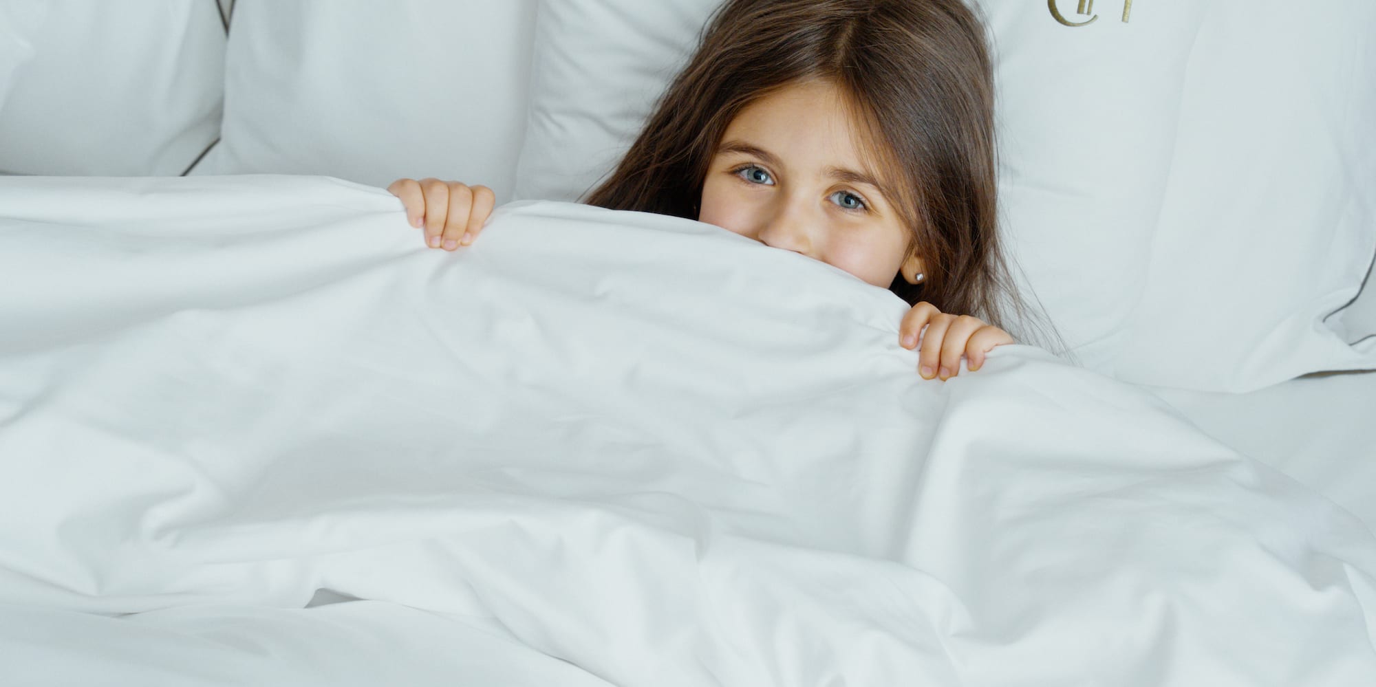 Young girl peeking playfully from under white bed covers with monogrammed pillows.