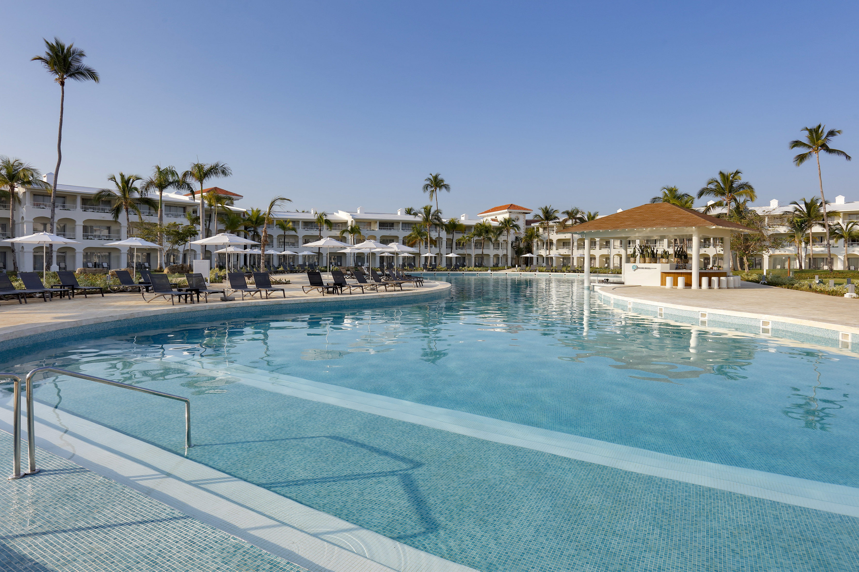 Tropical resort pool, swim-up bar, hotel buildings, and palm trees.