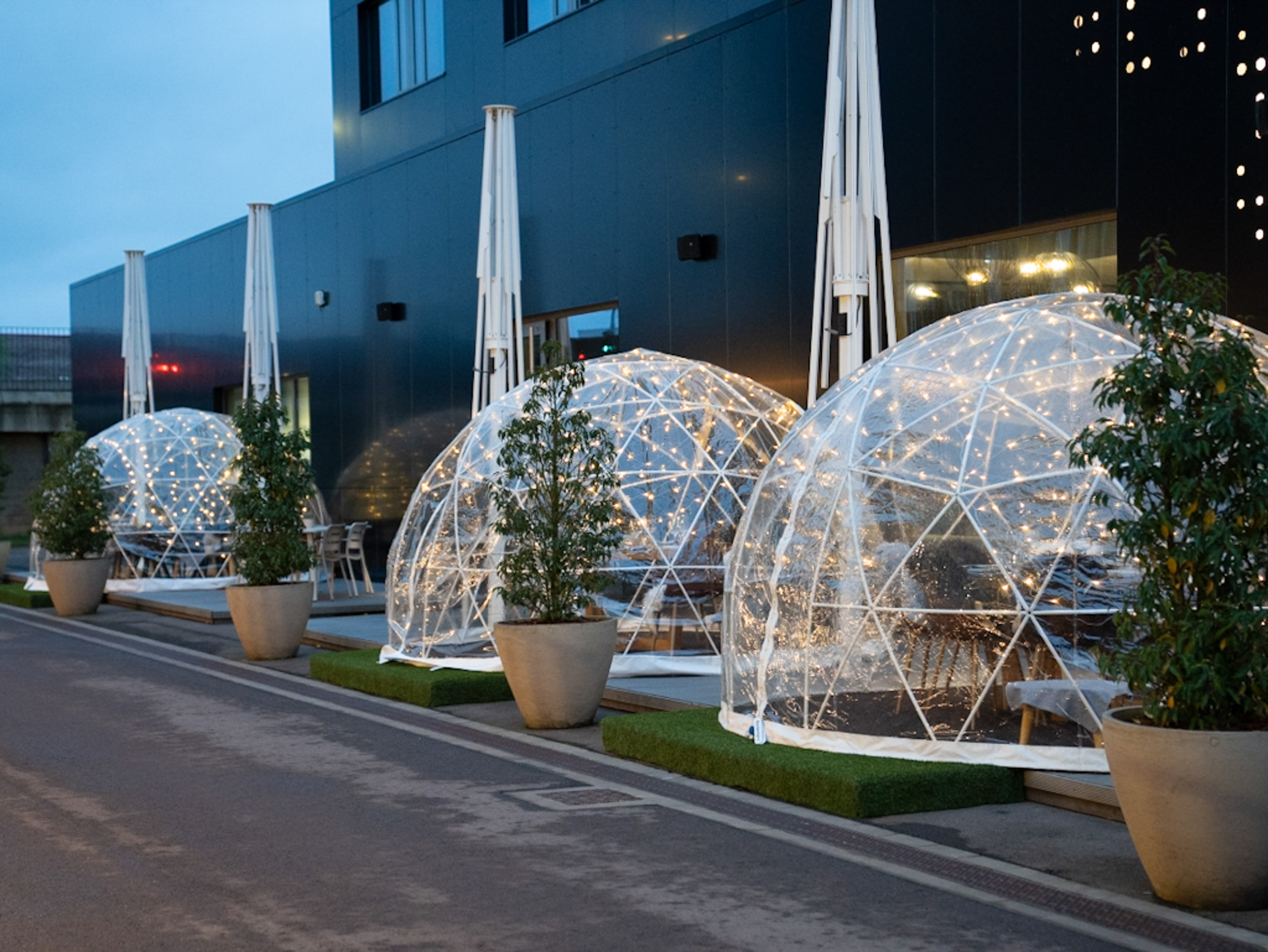 Geodesic dining igloos with string lights outside a dark modern building.