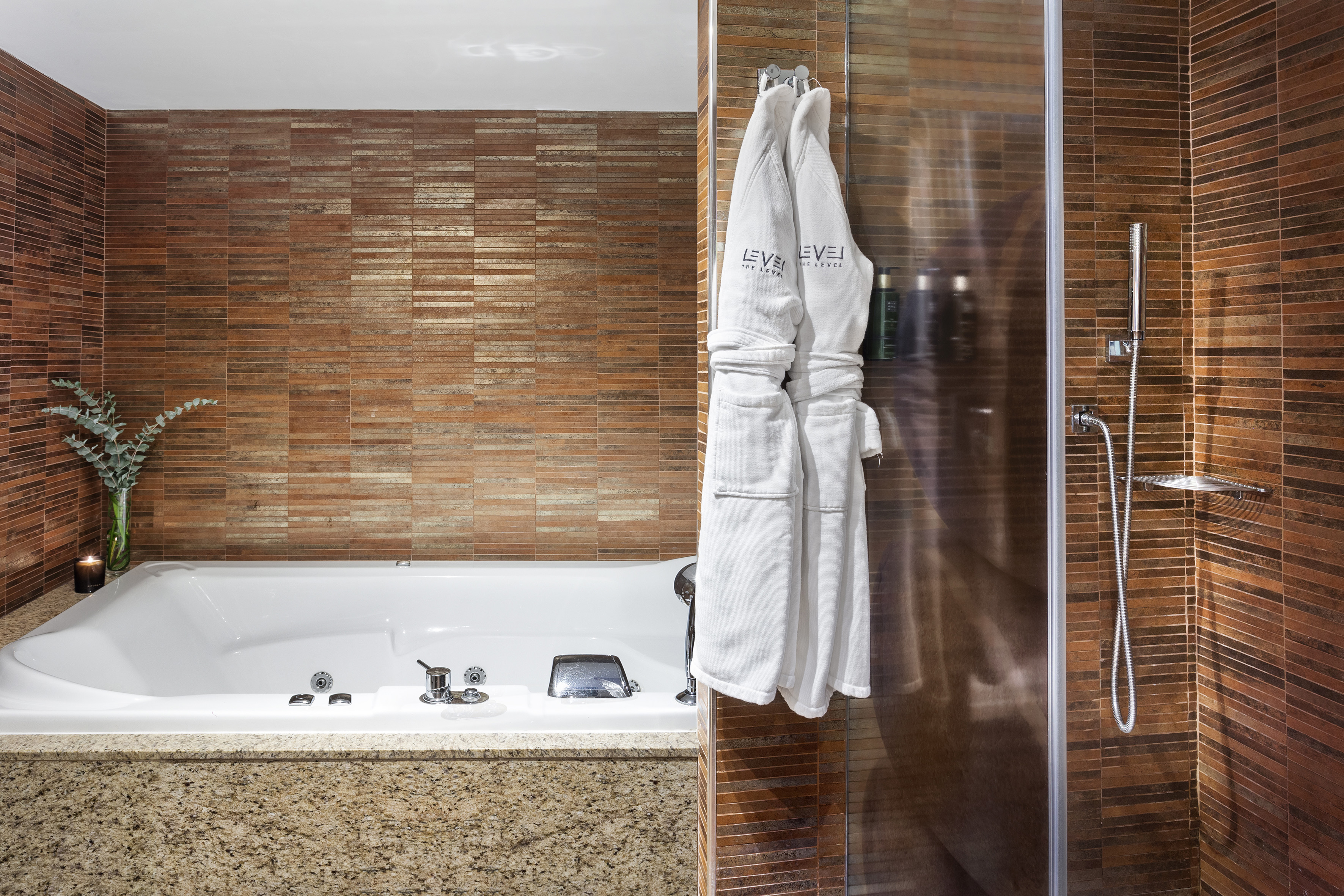 Hotel bathroom with white jacuzzi tub, glass shower, brown tiles, and LEVEL robes.