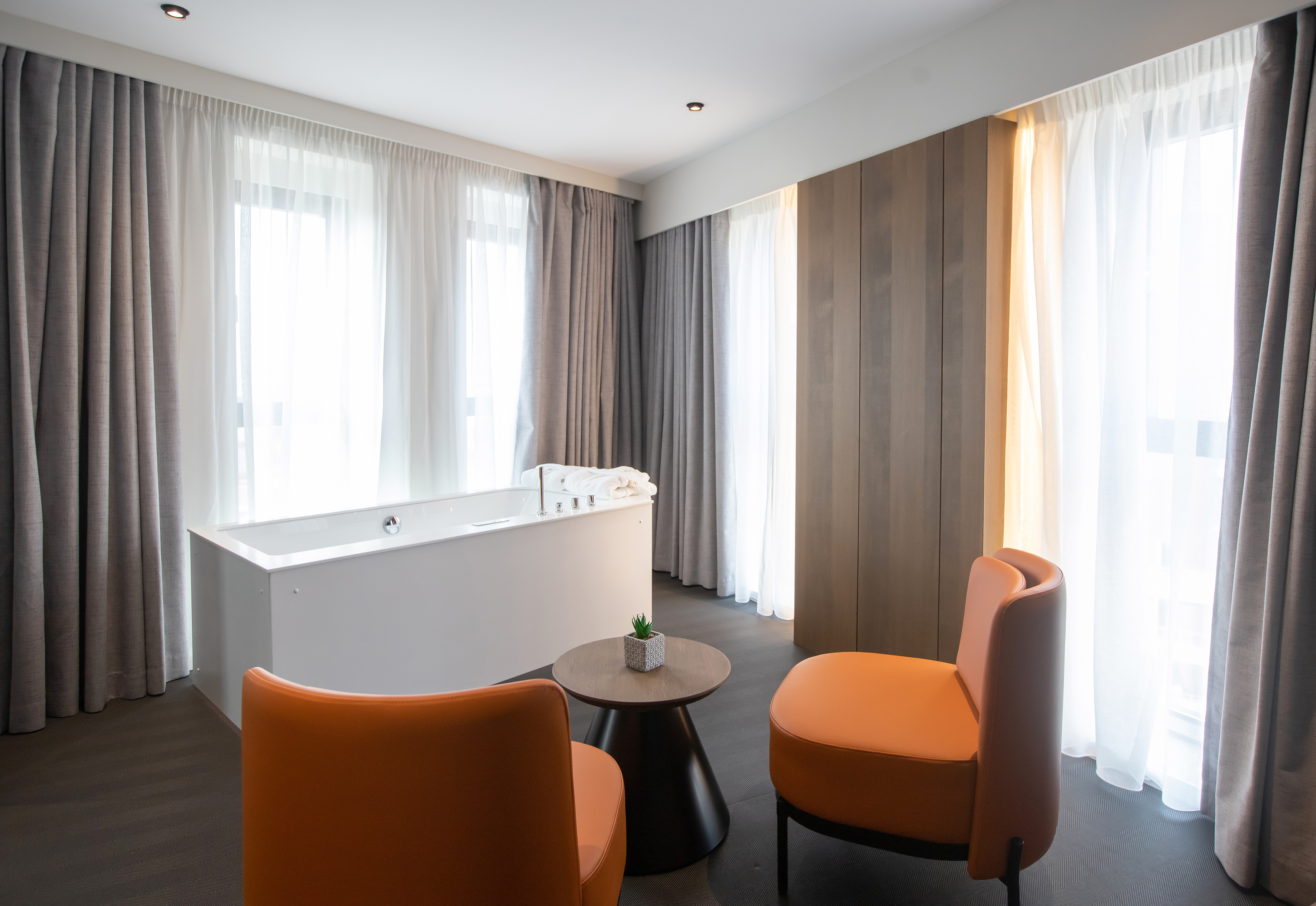 Modern bathroom suite with white tub, orange chairs, and bright windows.