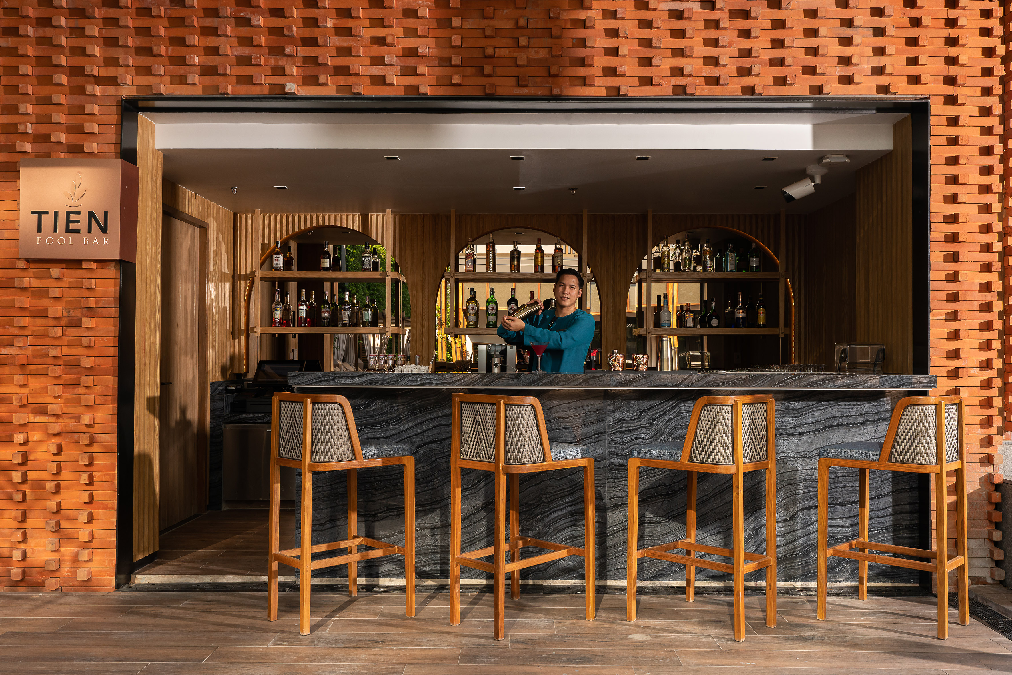 Bartender shaking cocktail at Tien Pool Bar with brick facade and stools.