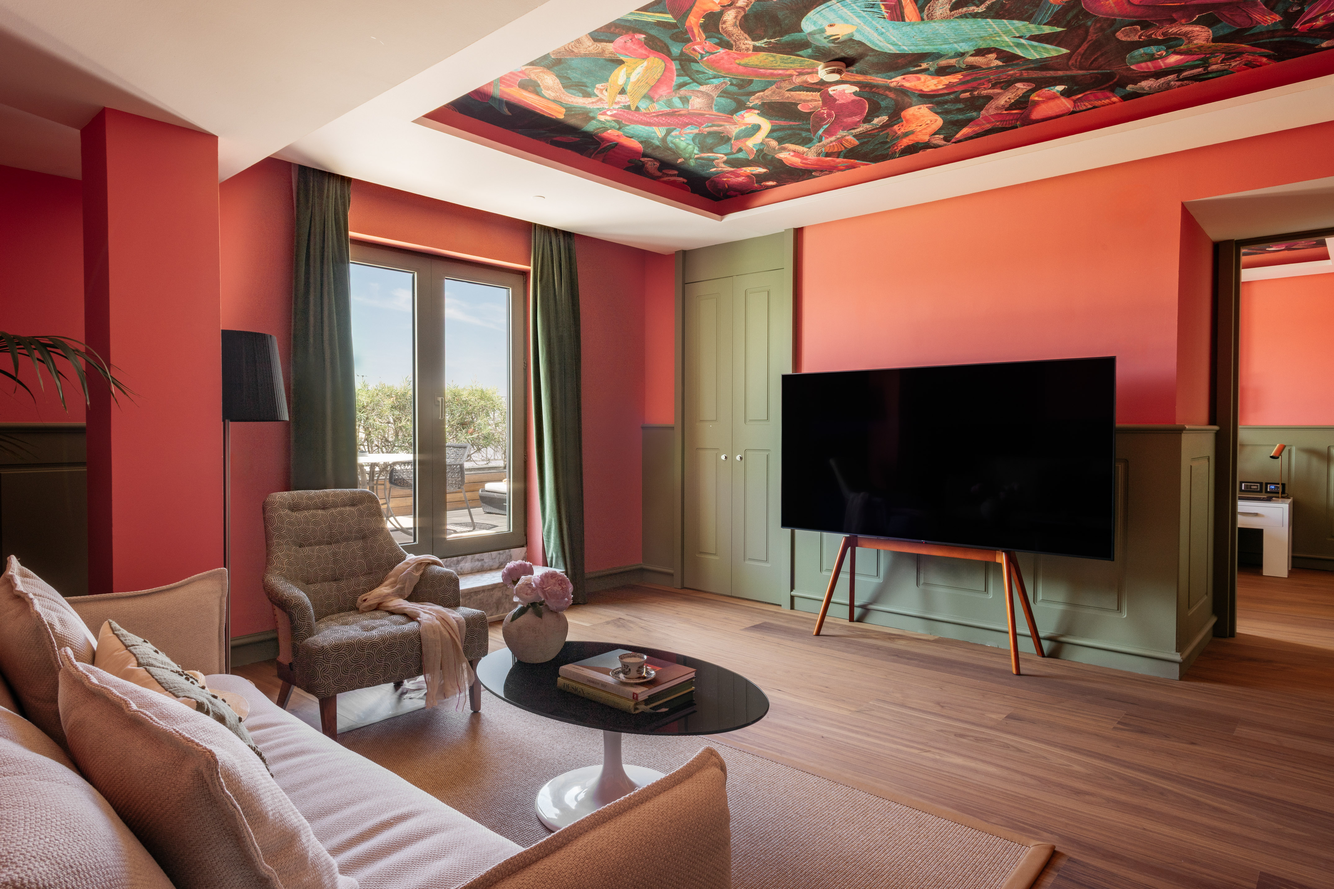Suite moderna coral y verde con techo mural de aves, TV, sofá y balcón.
