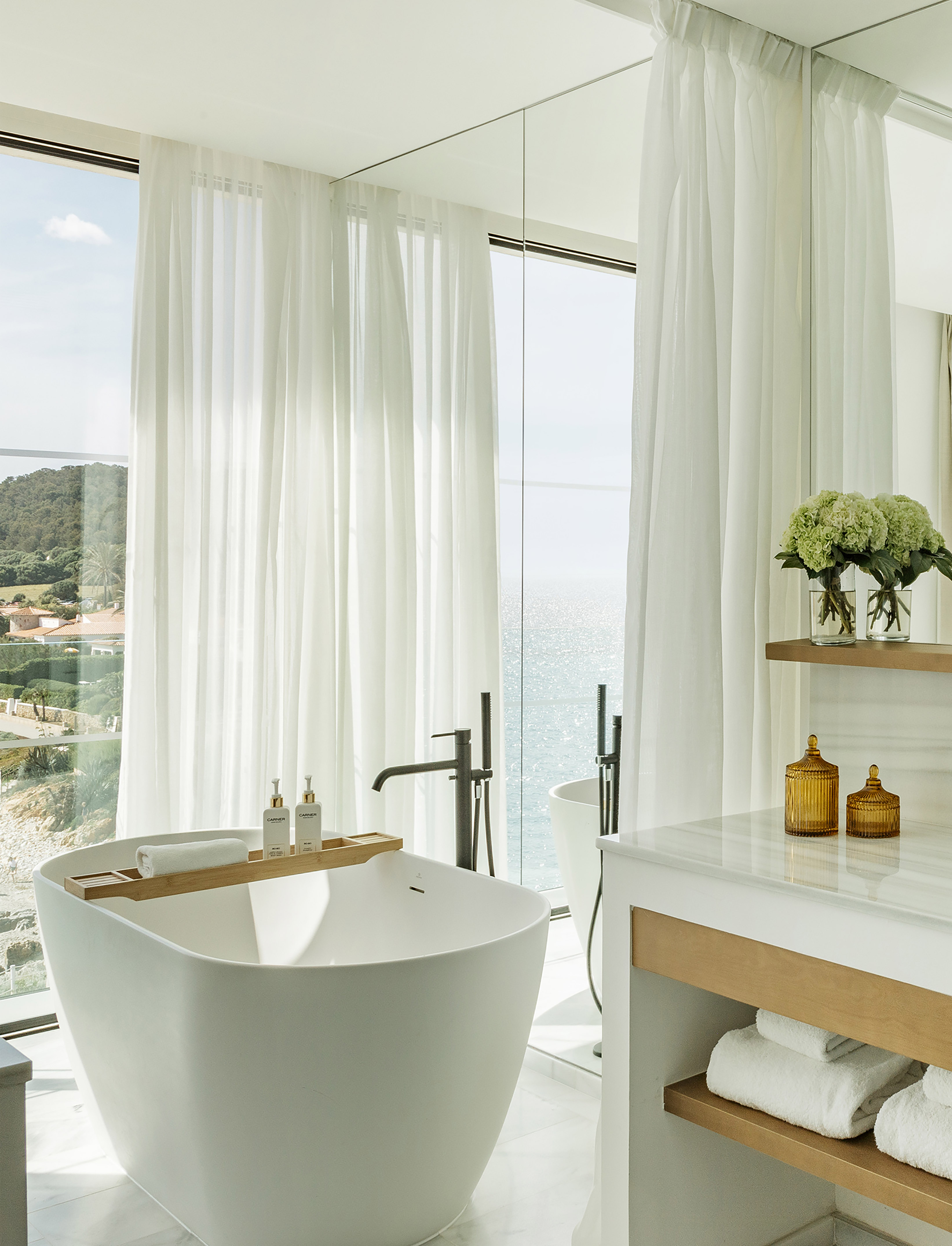 Modern bathroom with ocean view, white freestanding tub, sheer curtains.