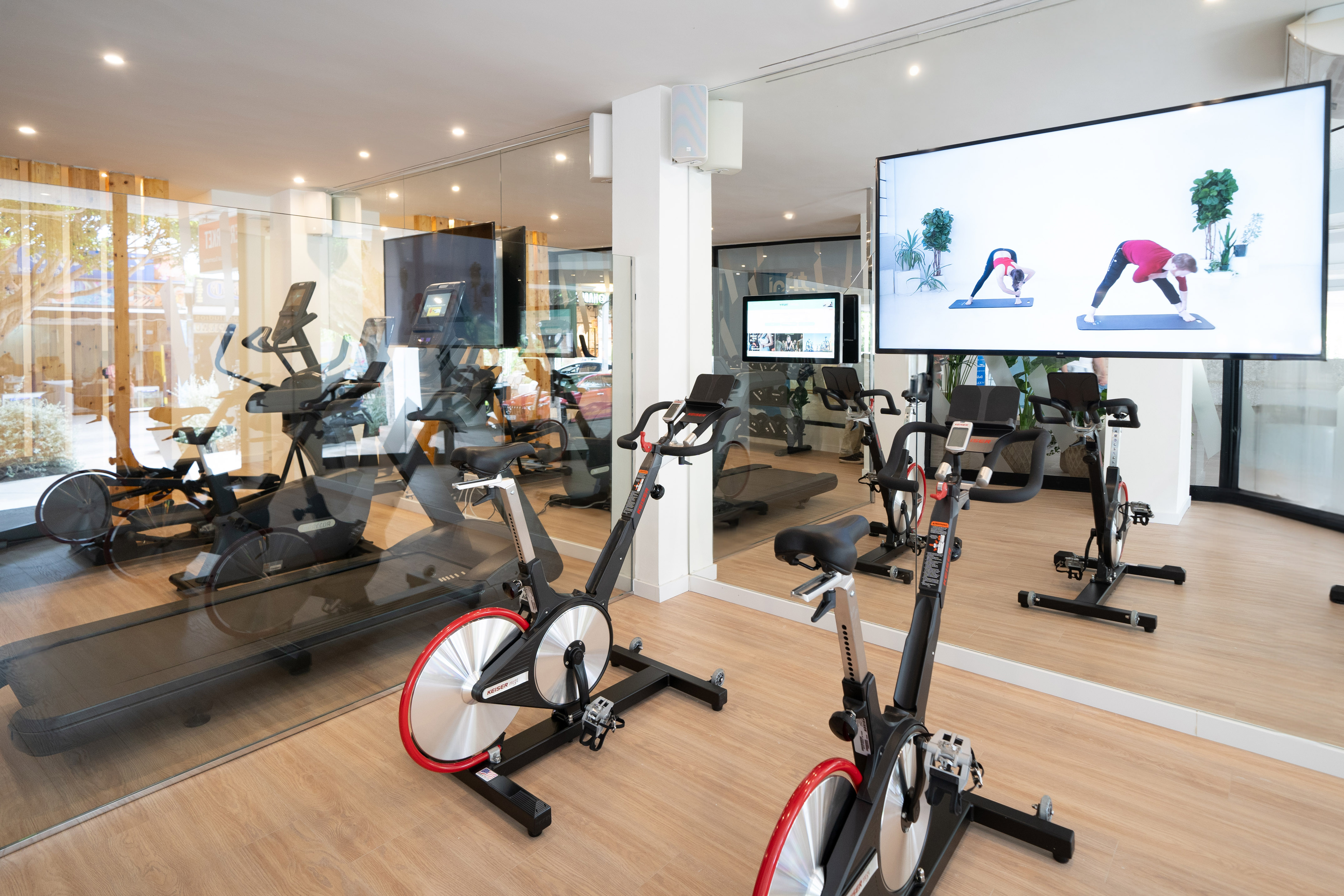 Bright fitness room with spin bikes, treadmills, and screen displaying a workout.