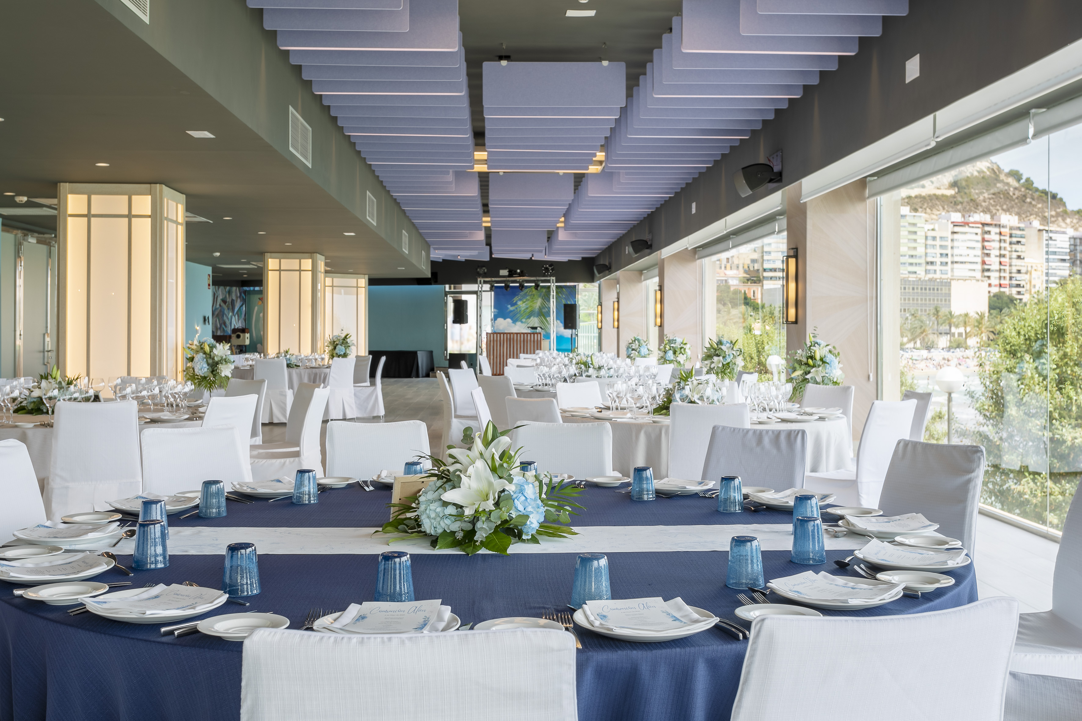 Elegant blue and white event hall, tables set with flowers, panoramic view.