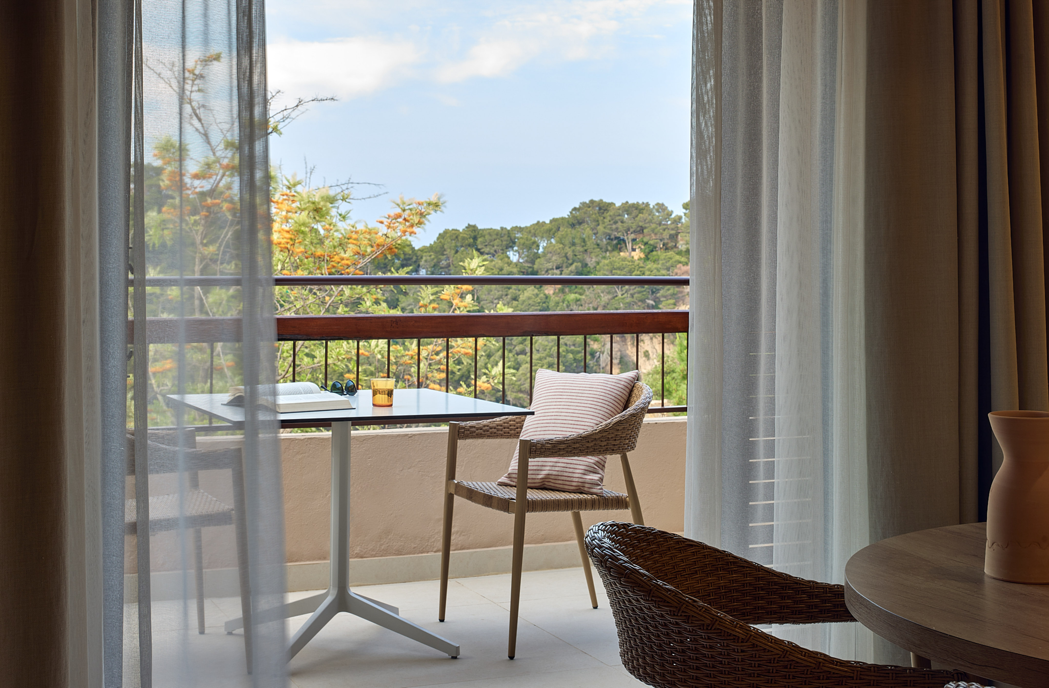 Balcony with outdoor table, chair, book, drink, and lush forest view.