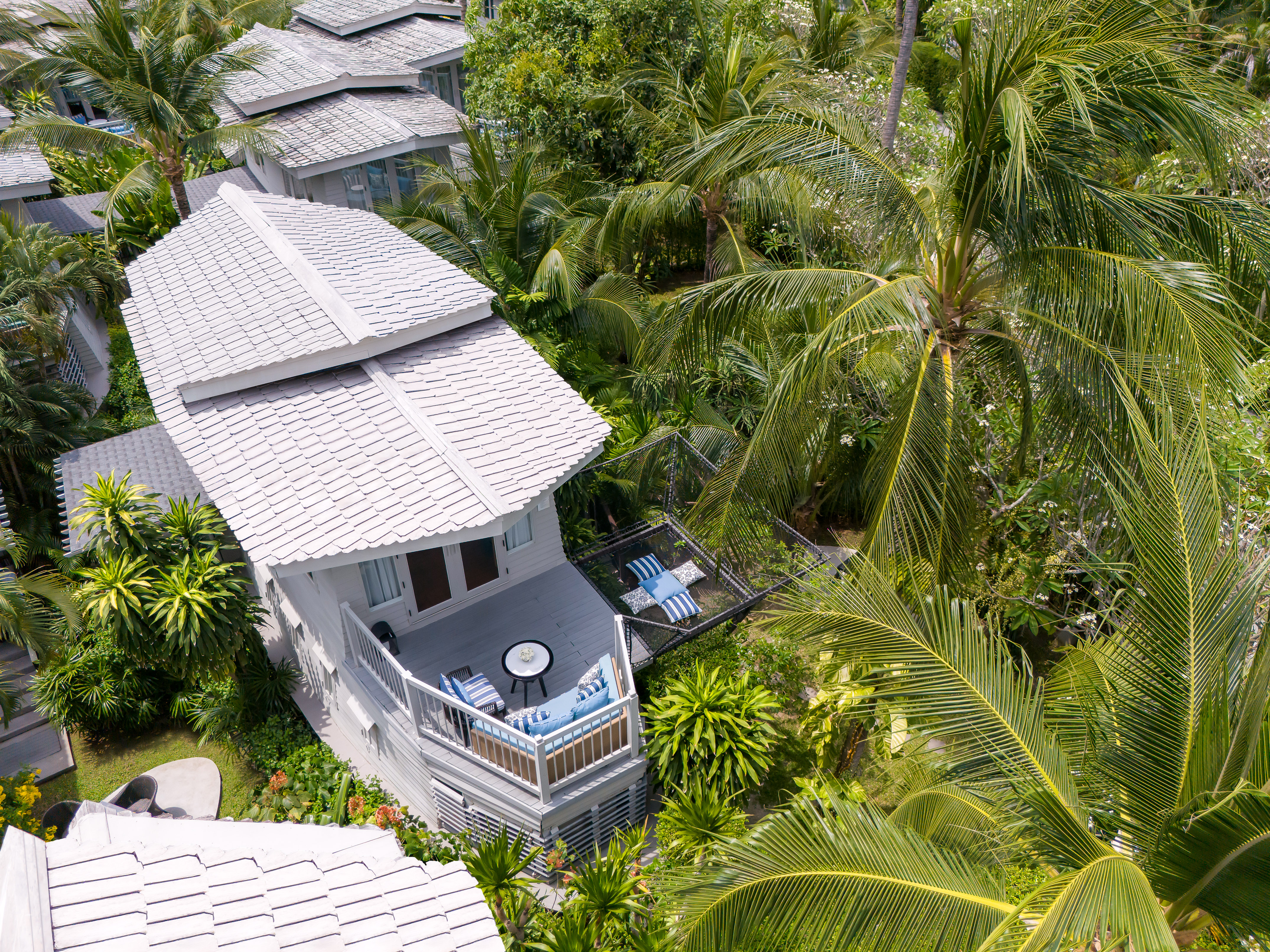 Tropical resort villa with white roof, balcony, hammock, and palm trees.
