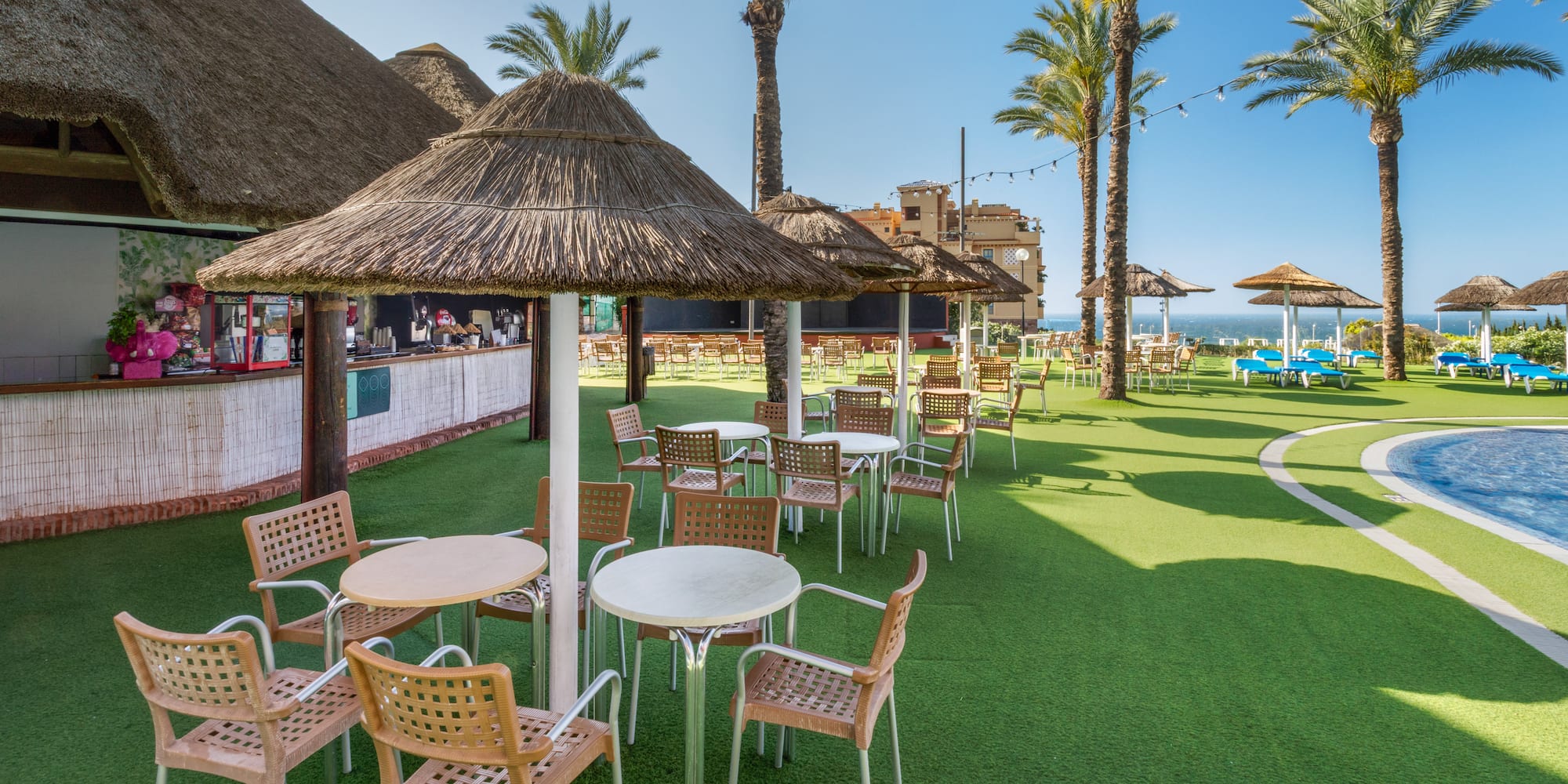 Resort poolside bar with thatched umbrellas, outdoor tables, chairs, and palm trees.