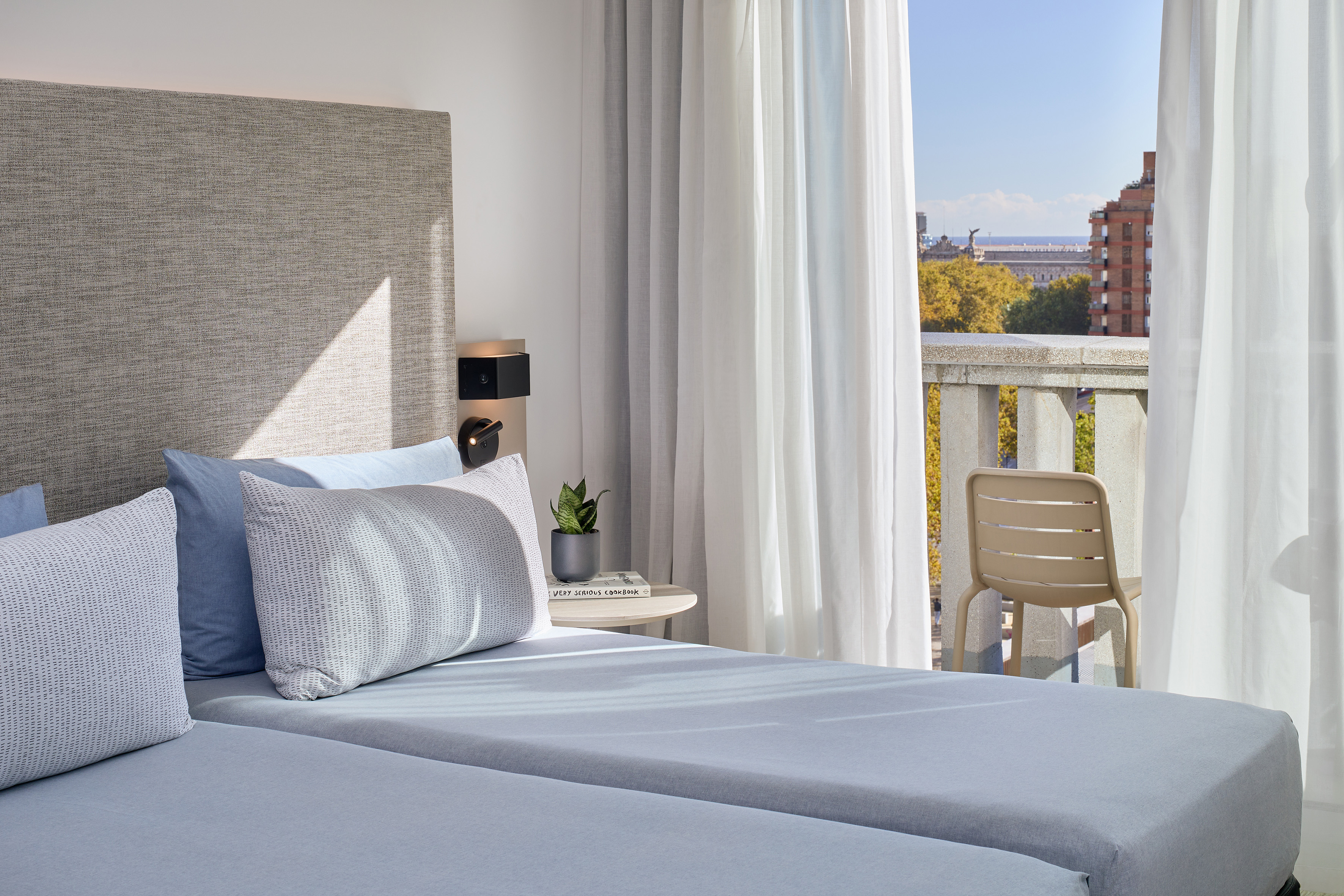 Modern hotel room with twin beds, light blue bedding, balcony, and city view.