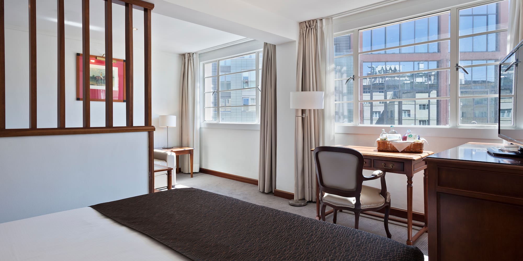 Bright hotel room with bed, wooden divider, desk, and large city view windows.