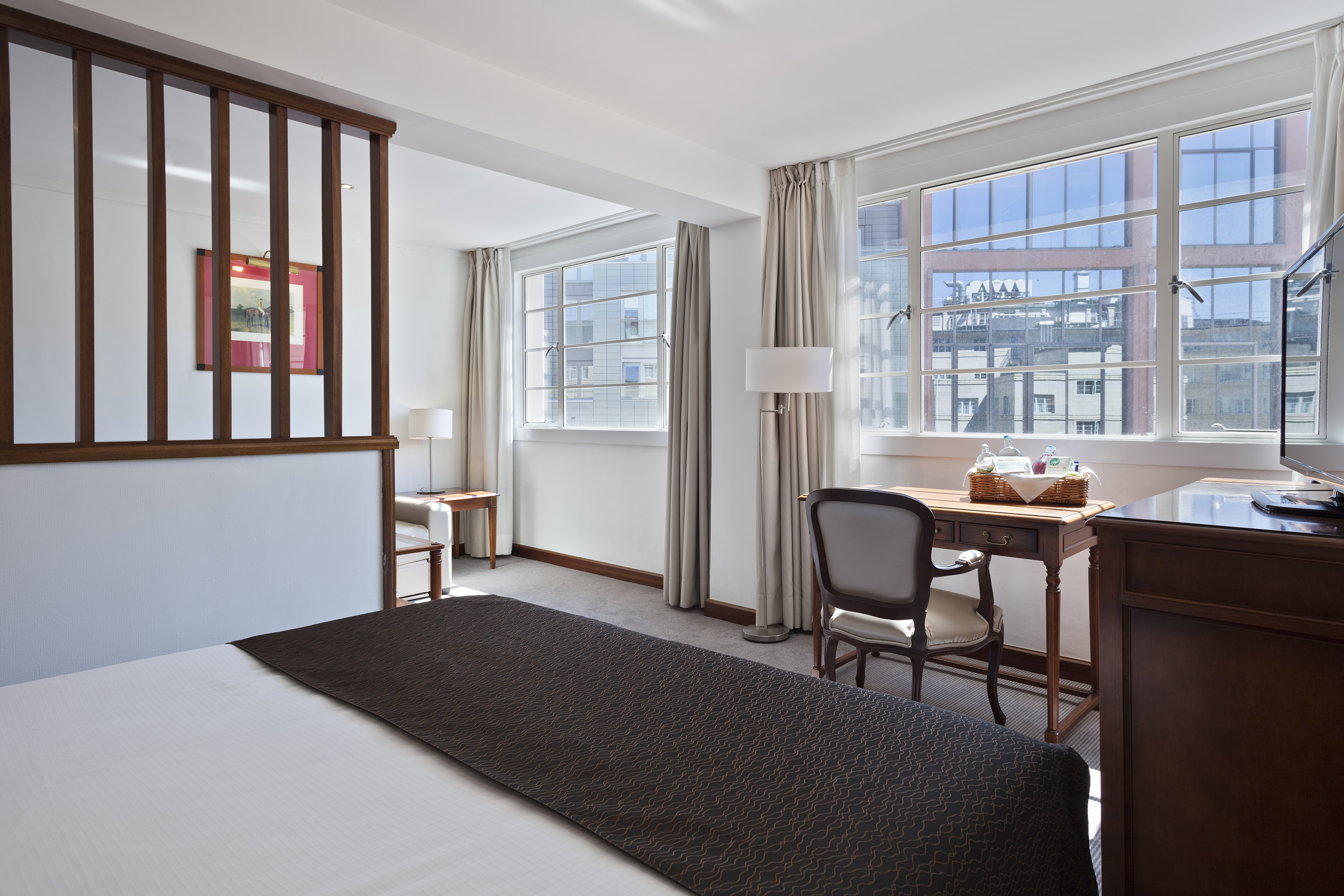 Bright hotel room with bed, wooden divider, desk, and large city view windows.