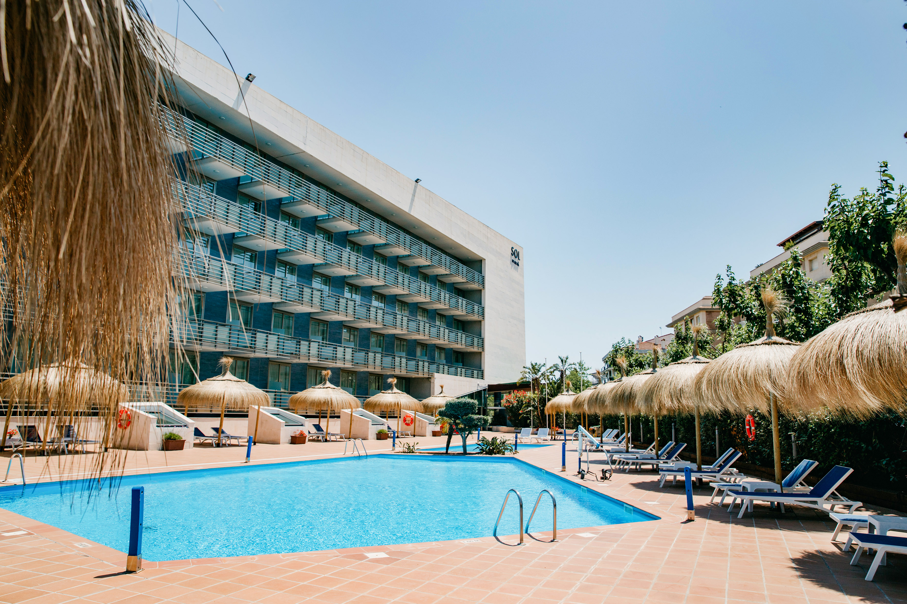 Sunny resort pool with straw umbrellas, loungers, and modern hotel building.