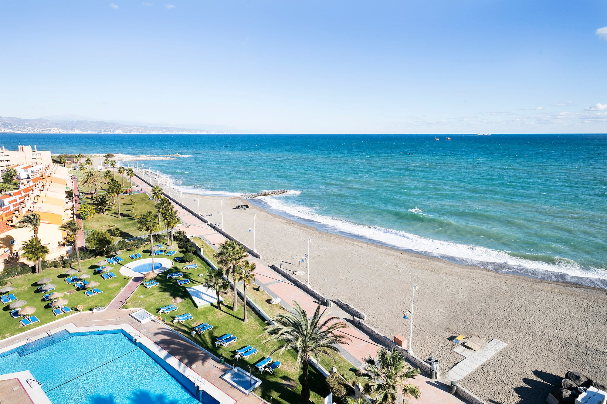 Aerial view of resort pool, palm trees, sandy beach, and blue ocean.