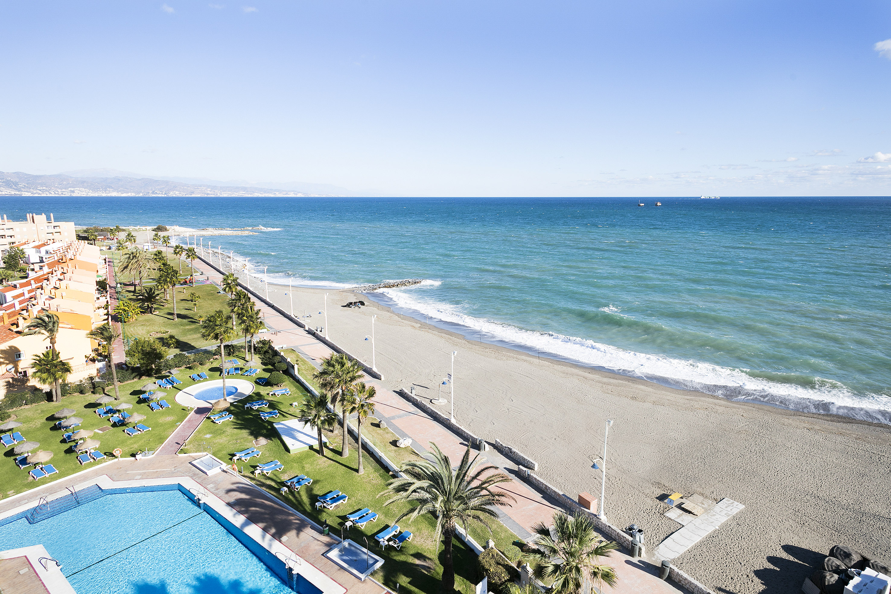 Aerial view of resort pool, palm trees, sandy beach, and blue ocean.