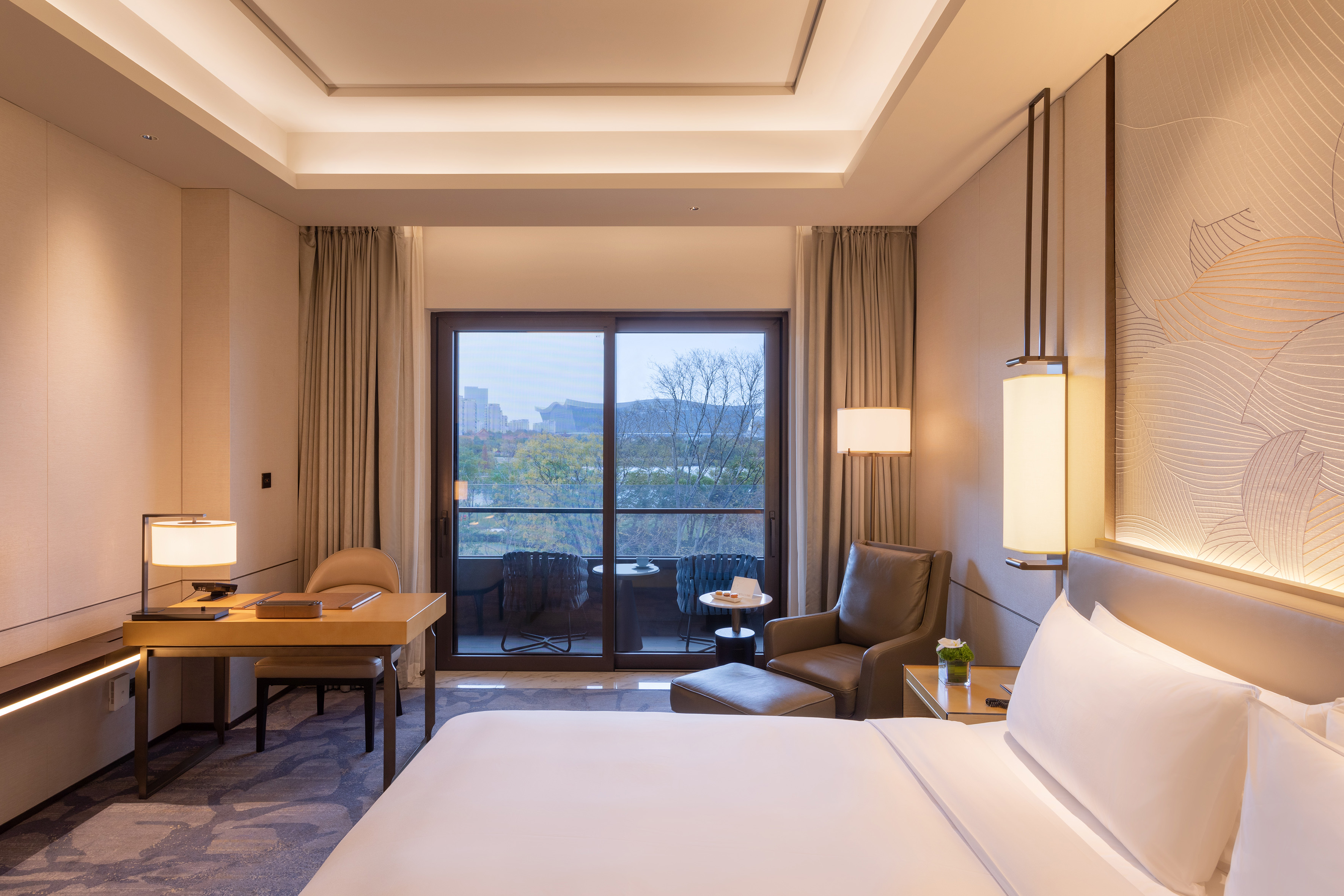 Modern luxury hotel bedroom with a balcony, desk, and urban landscape view.