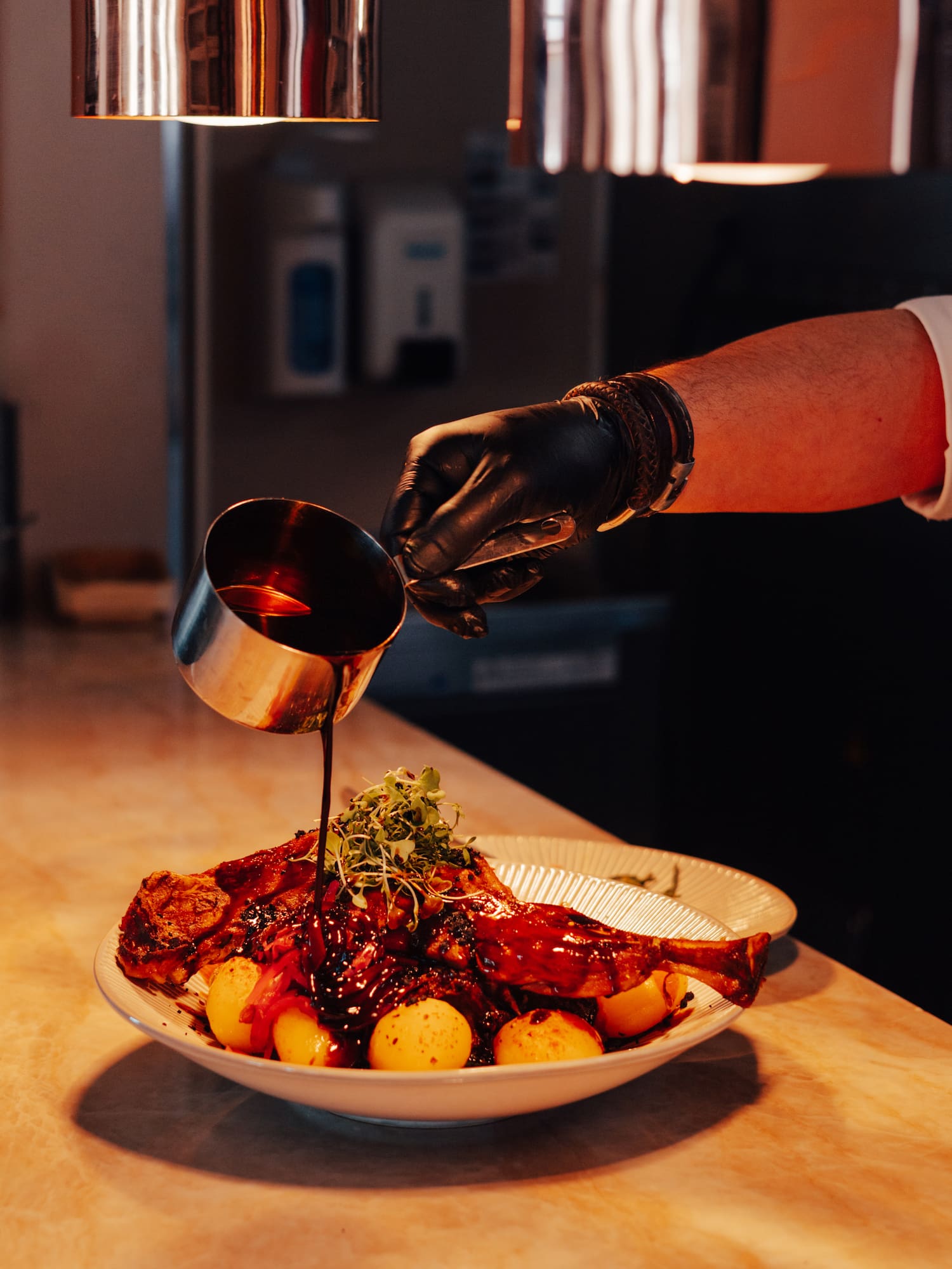 Chef's gloved hand pours dark sauce over roasted meat, potatoes, and sprouts.