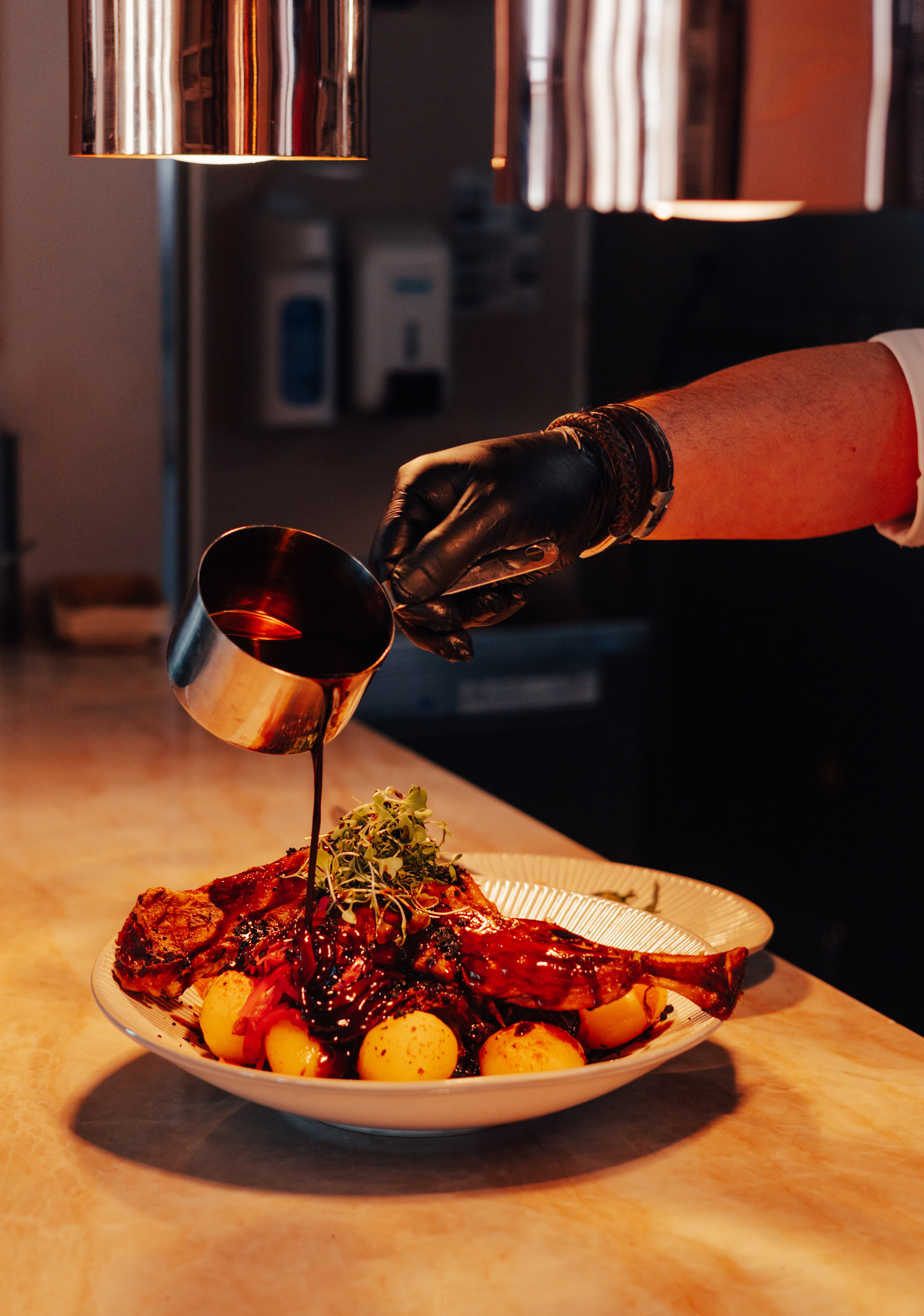 Chef's gloved hand pours dark sauce over roasted meat, potatoes, and sprouts.