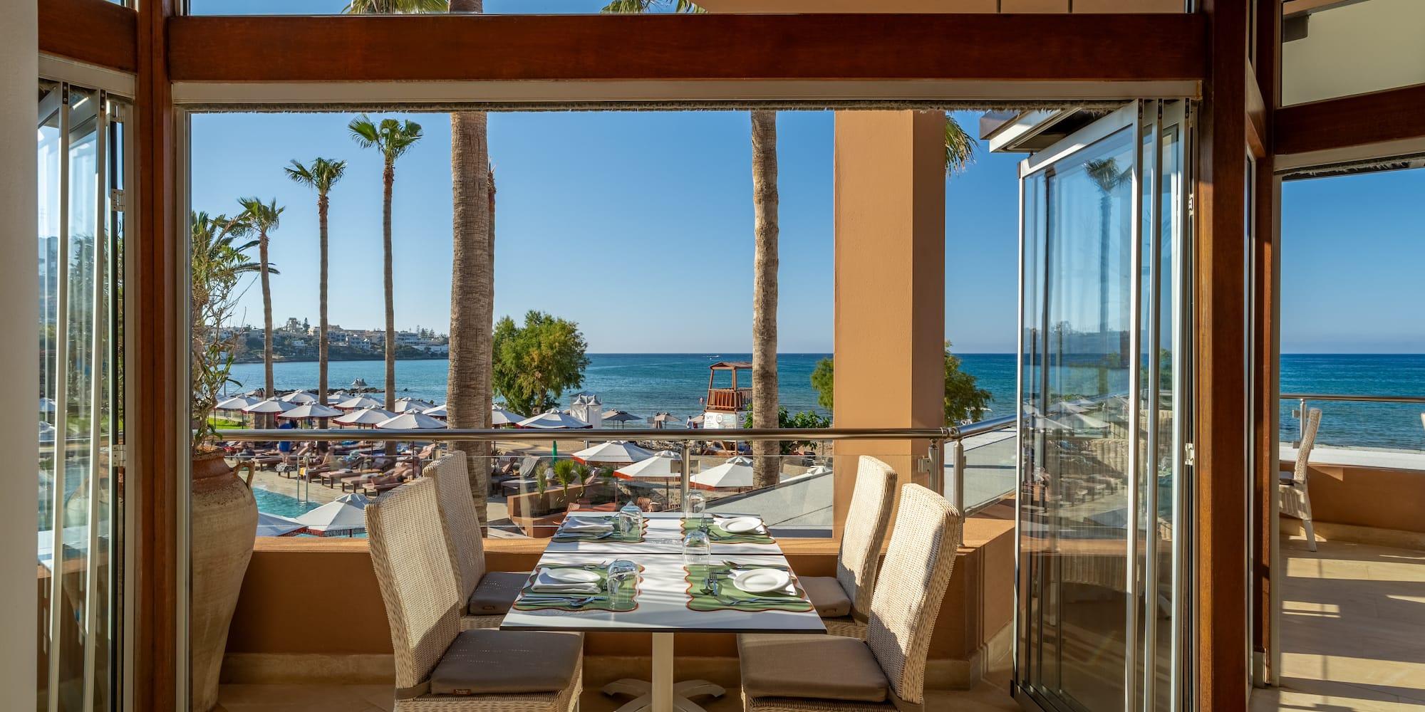 Restaurant balcony with sea view, palm trees, beach, and pool.