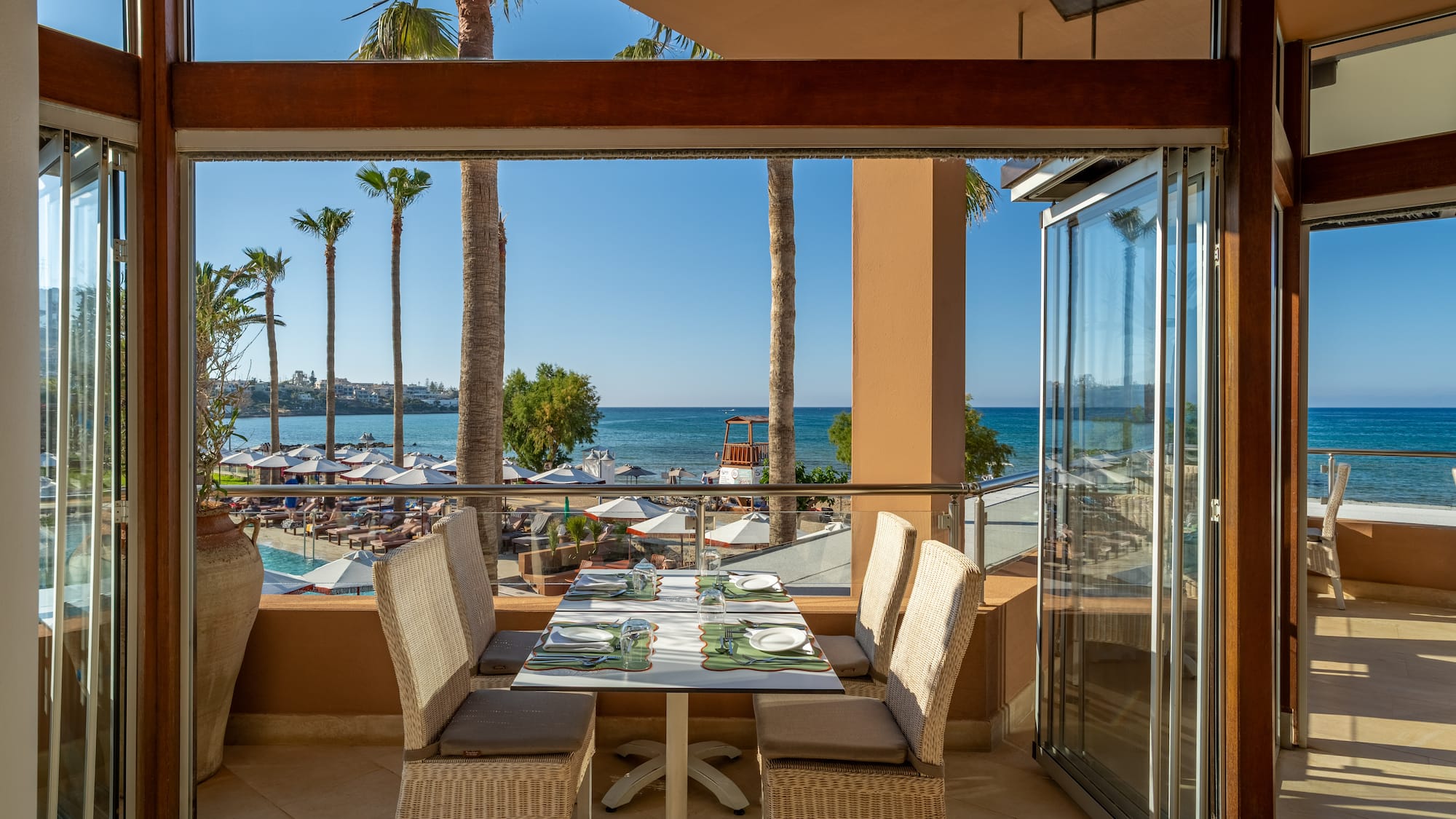 Restaurant balcony with sea view, palm trees, beach, and pool.