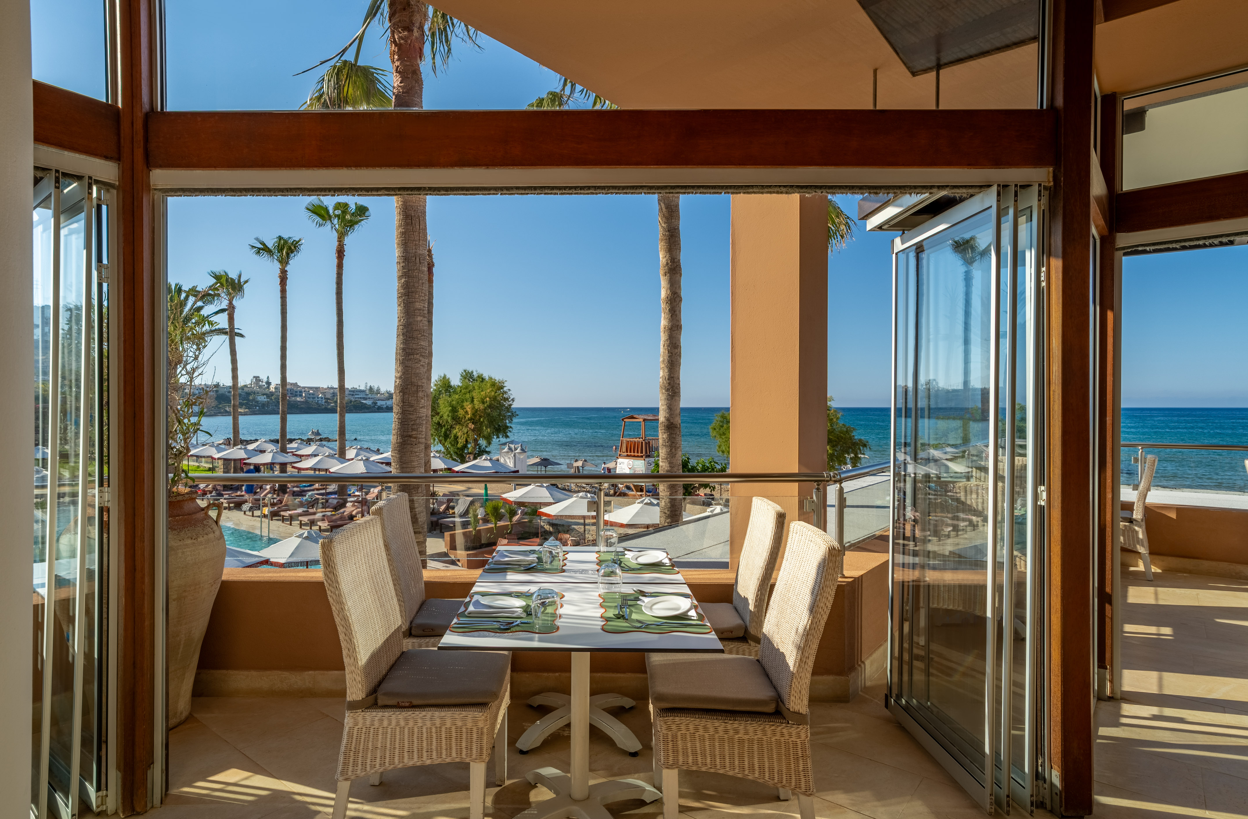 Restaurant balcony with sea view, palm trees, beach, and pool.