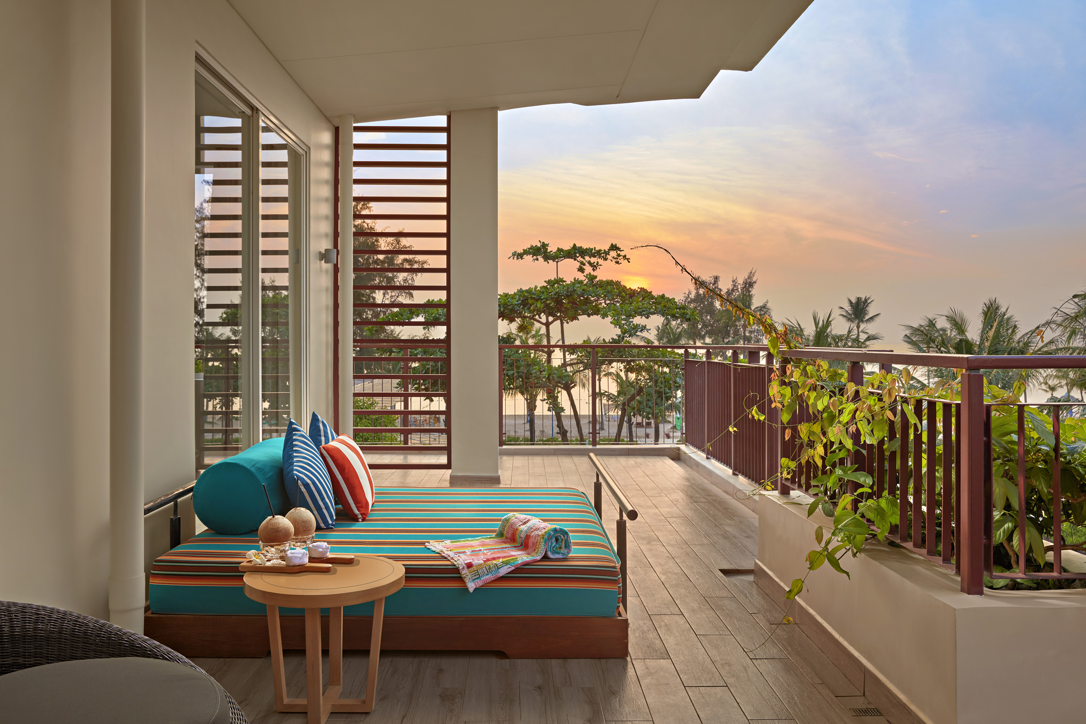 Tropical balcony daybed with colorful pillows, coconuts, and sunset beach view.