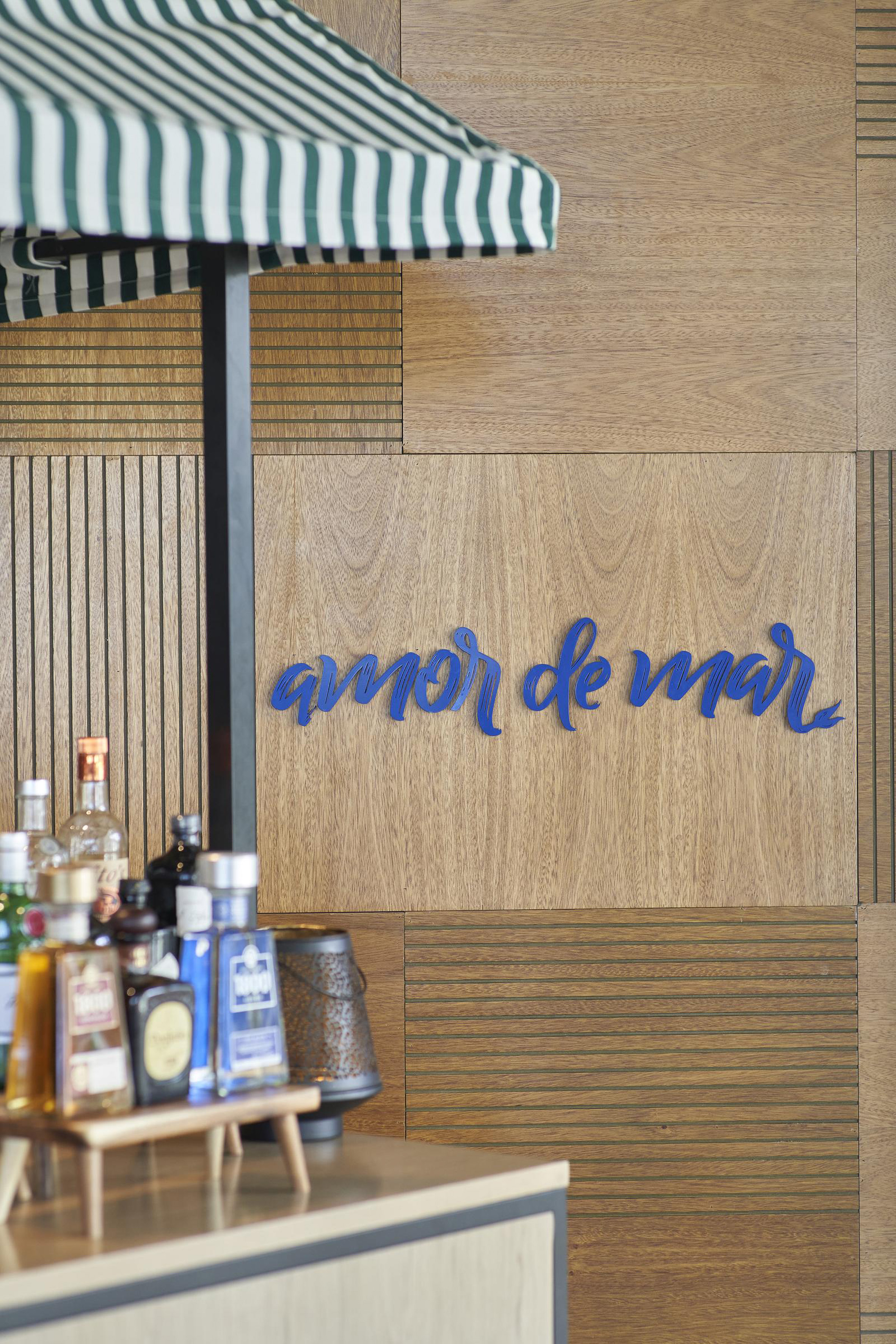 Amor de Mar sign on wood wall, bar with bottles, green striped awning.