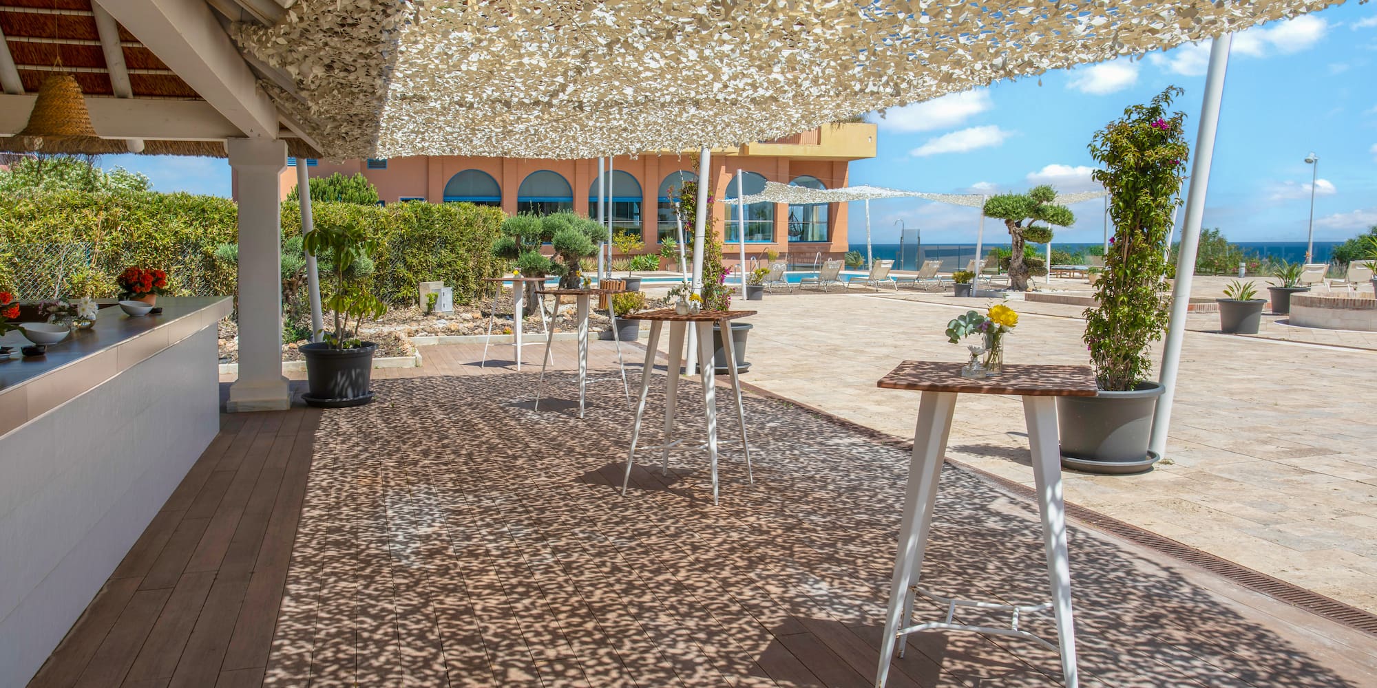 Resort patio with shade canopy, cocktail tables, bar, pool, and ocean view.