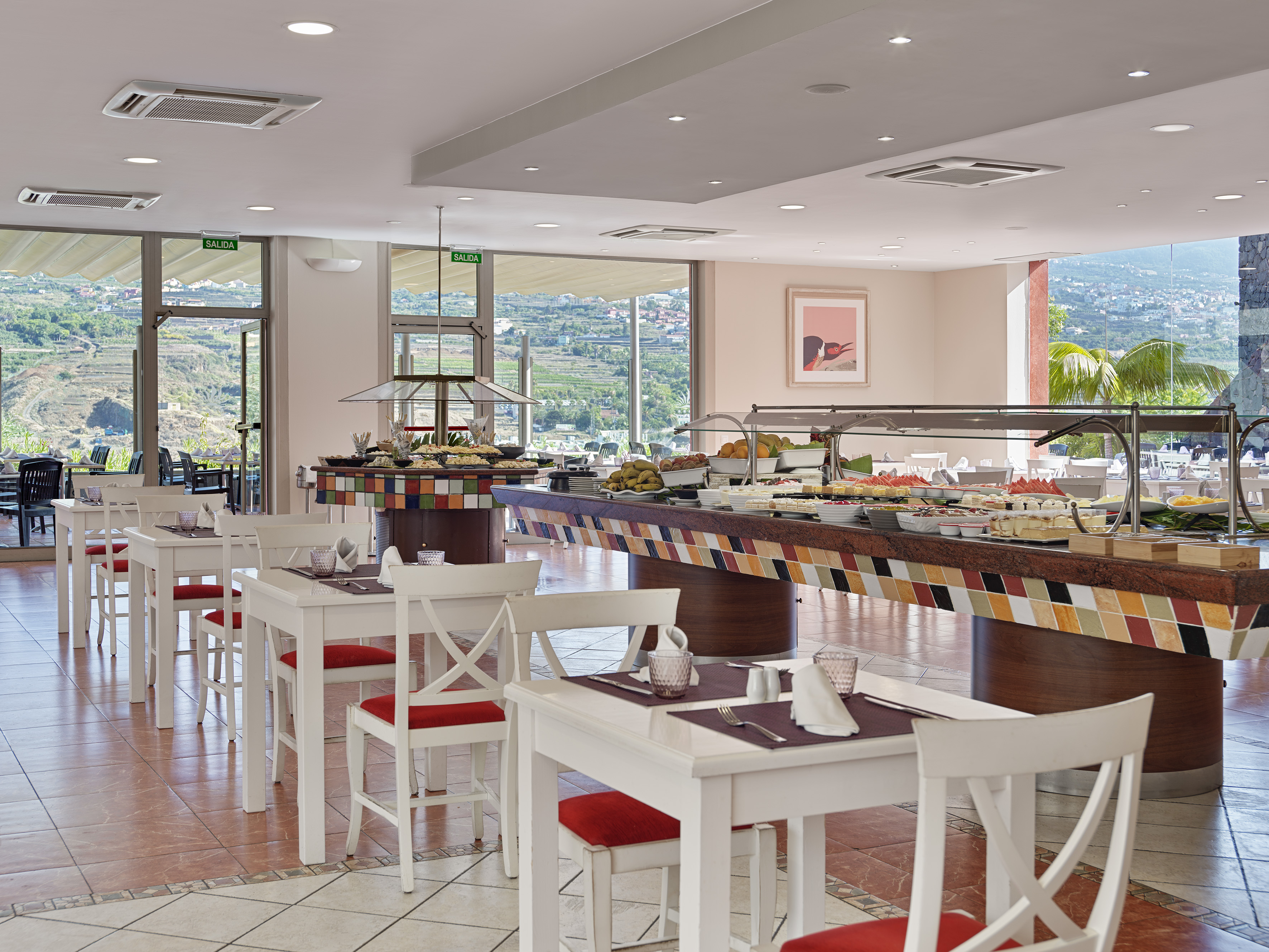 Restaurant buffet with varied dishes, white tables, red chairs, and scenic valley view.