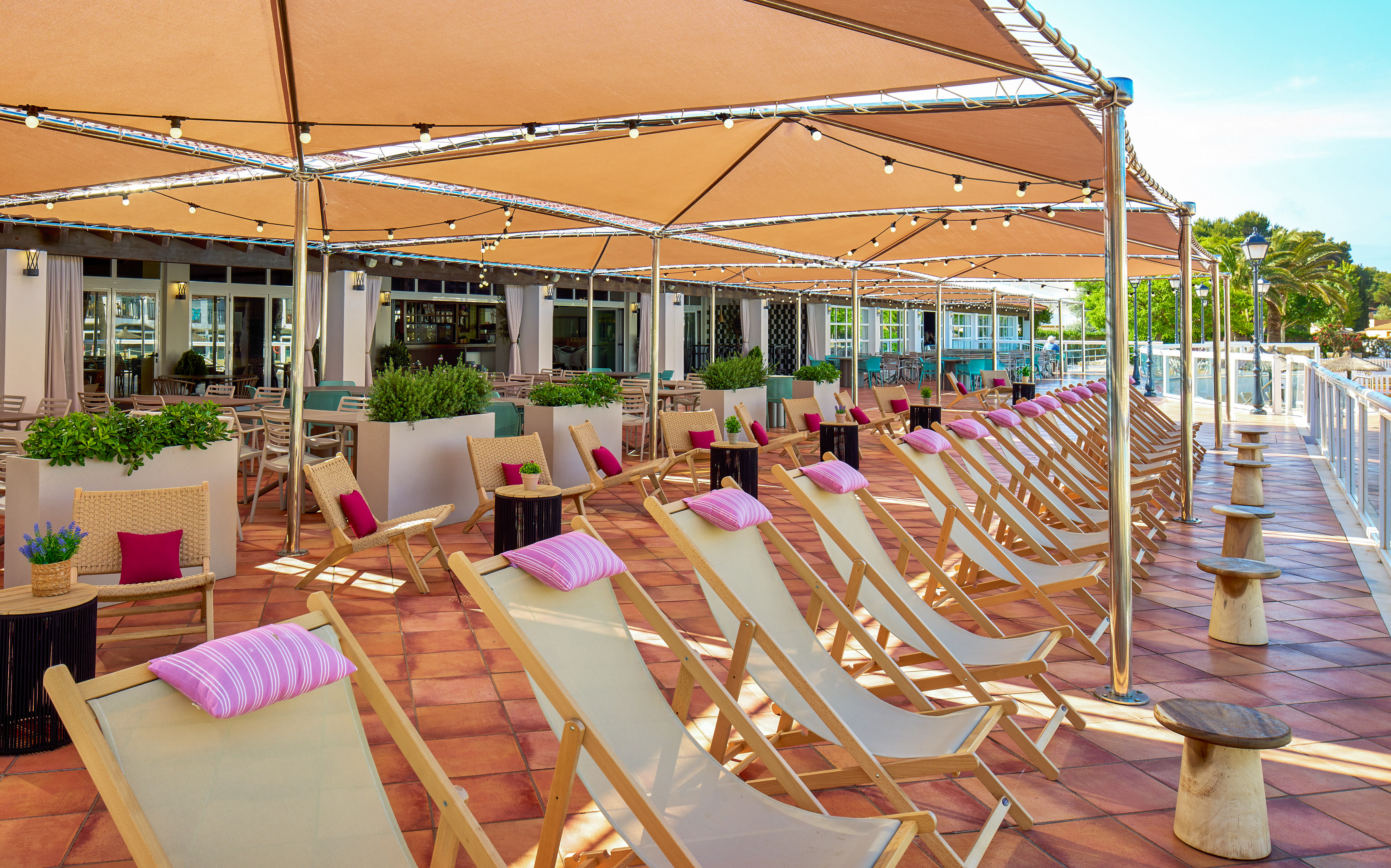 Shaded resort patio with rows of sun loungers, pink pillows, and string lights.