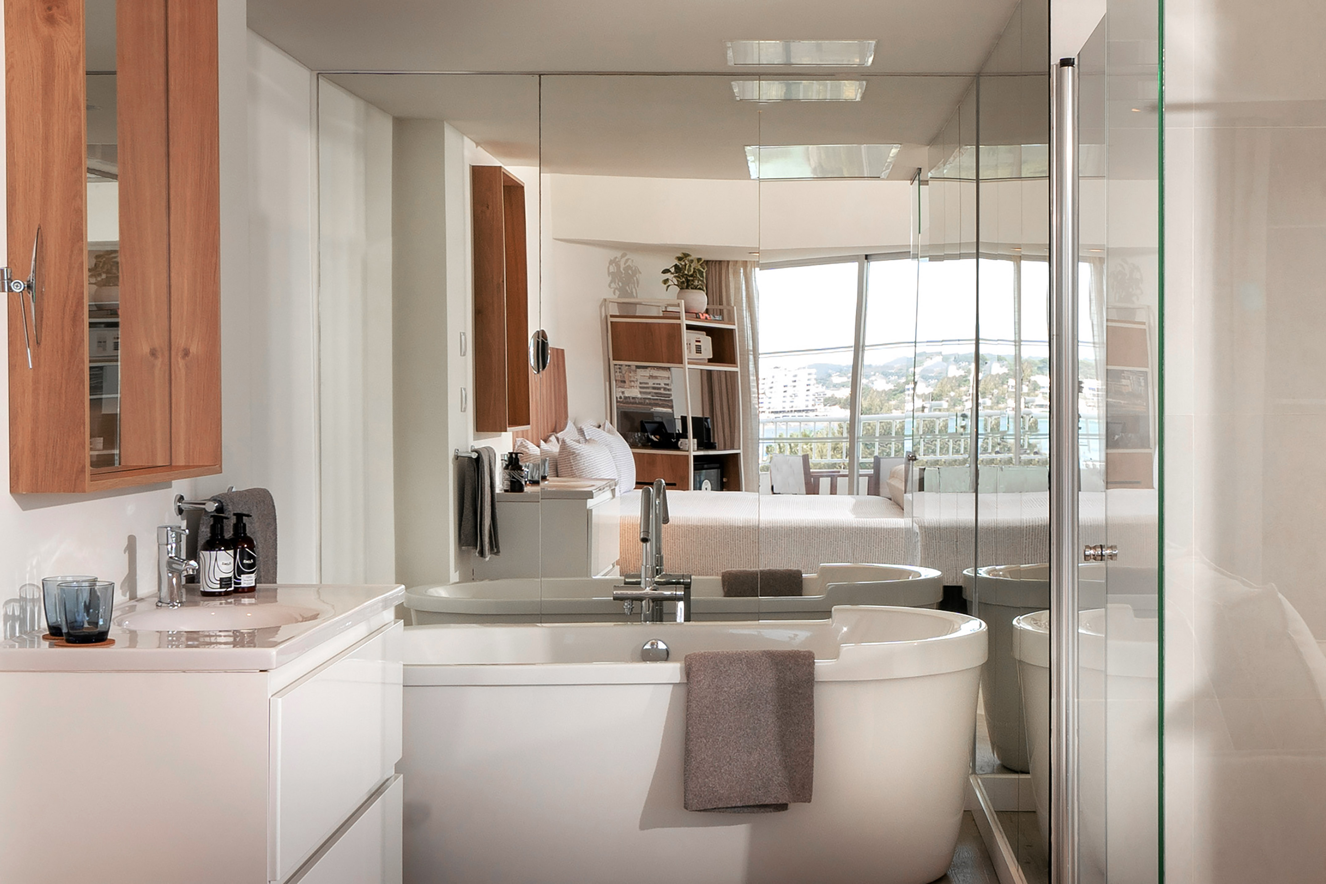 Modern bathroom with tub and vanity, reflecting bedroom suite and city view.