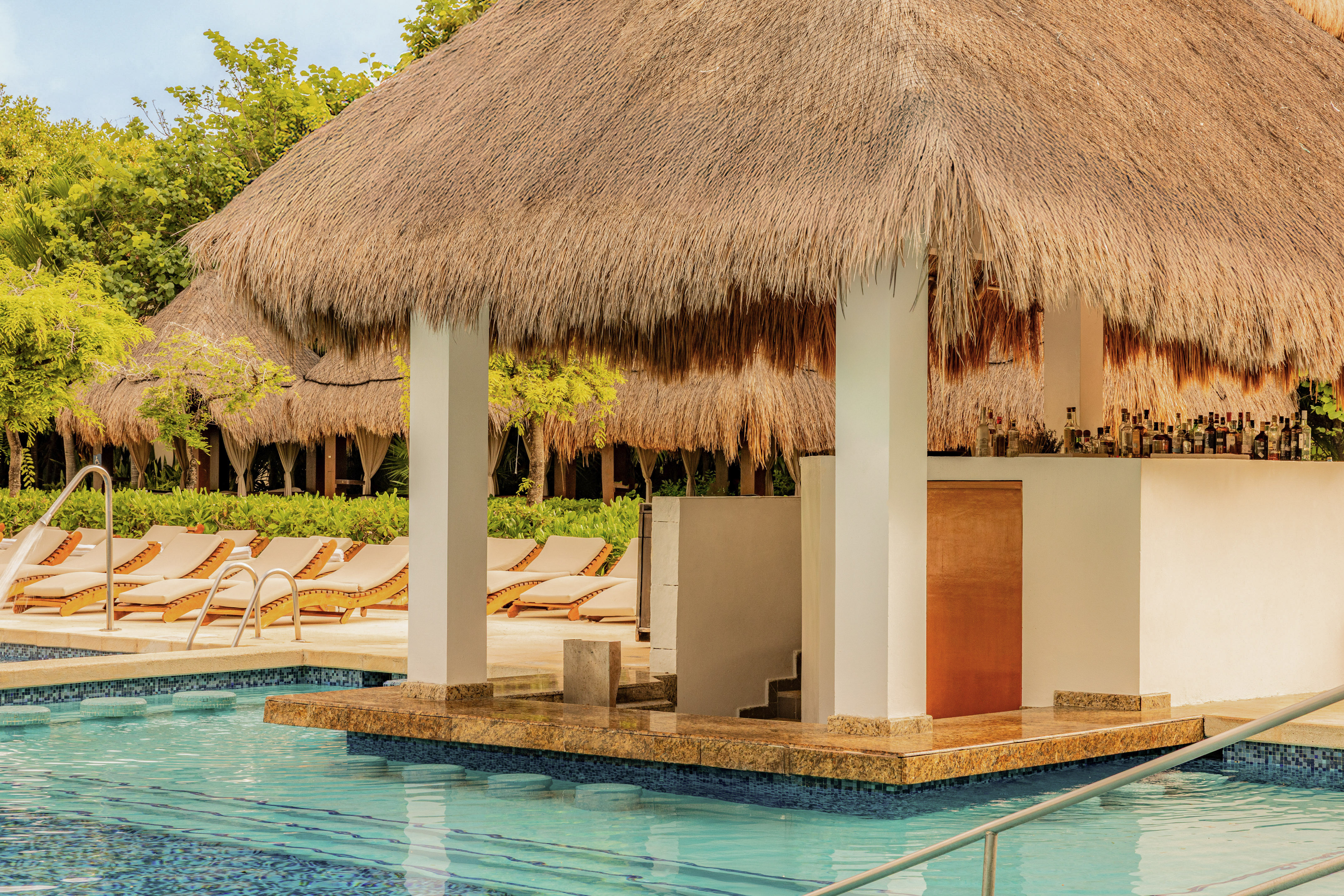 Thatched-roof swim-up bar with bottles by a tropical resort pool.