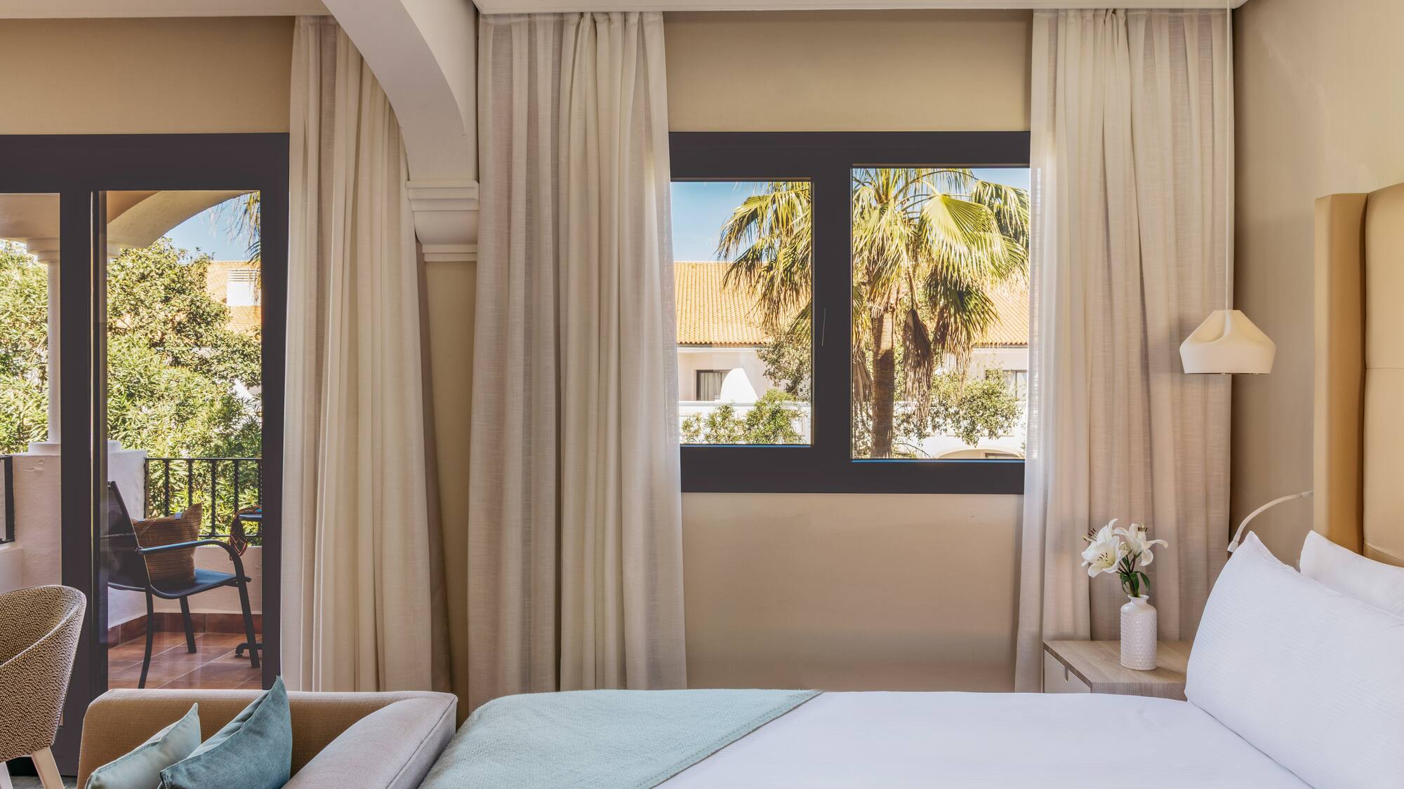 Hotel room with bed, sofa, balcony, and tropical palm tree view.