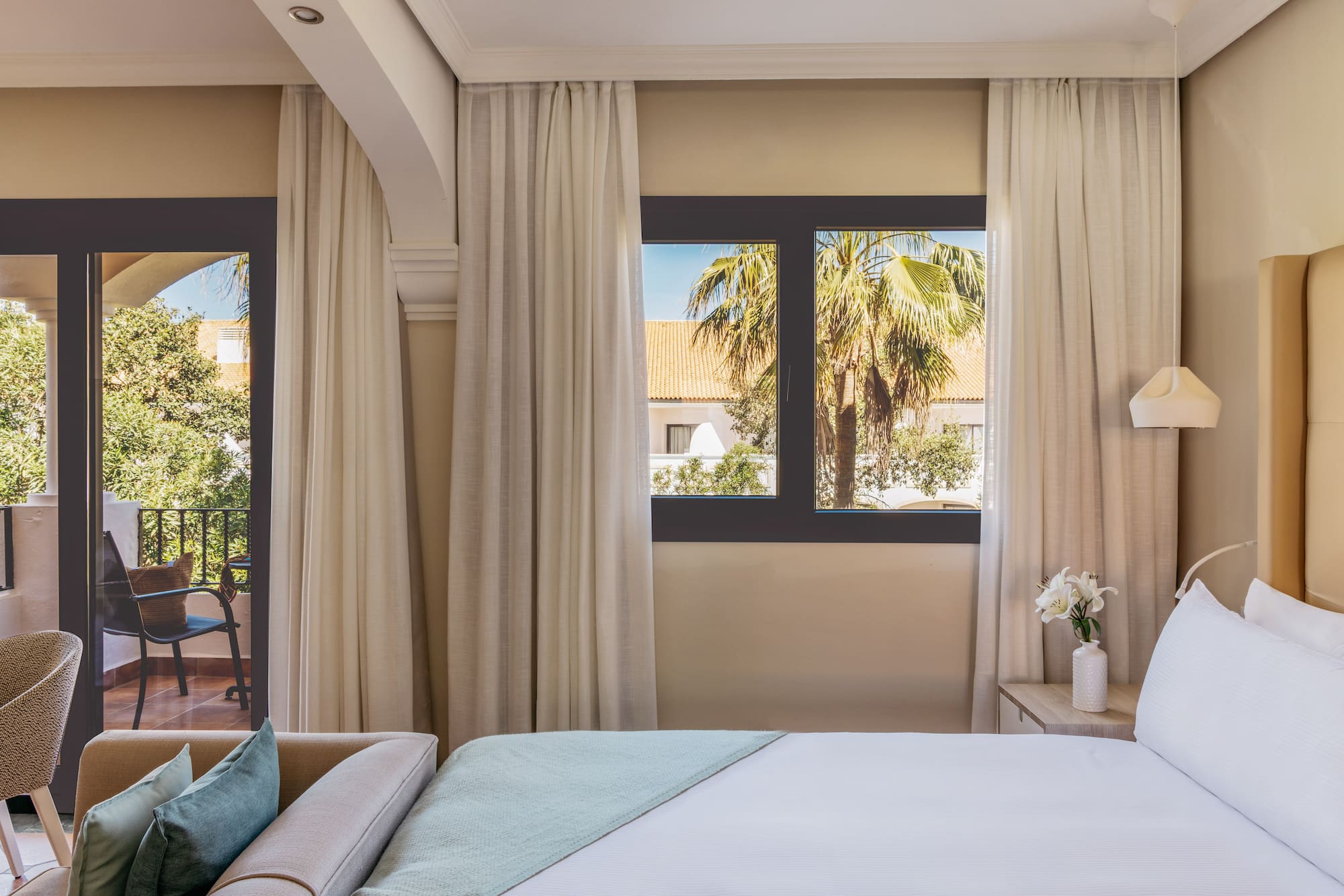 Hotel room with bed, sofa, balcony, and tropical palm tree view.