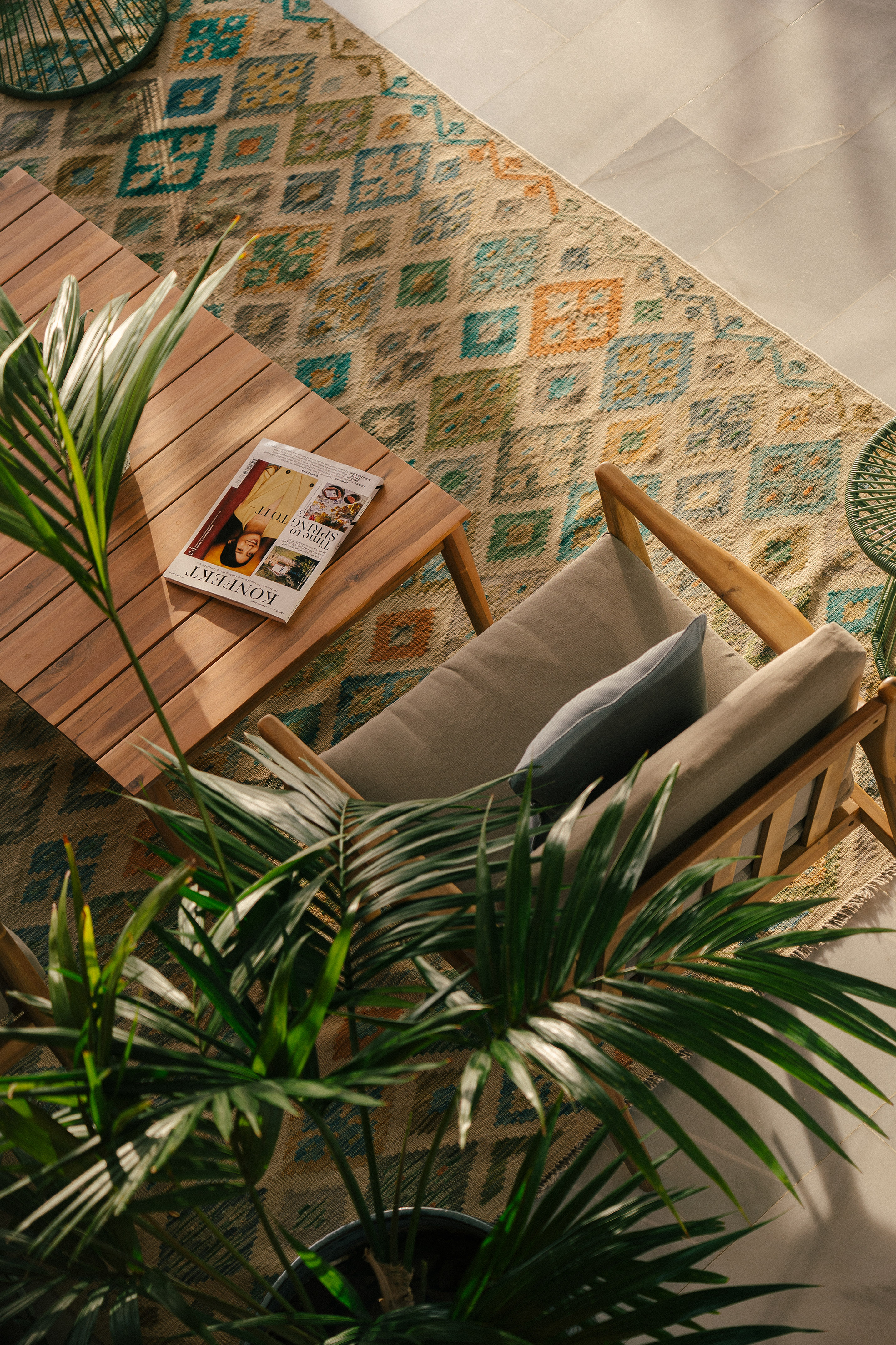 Outdoor wood furniture, patterned rug, magazine, and potted palm in sunlight.