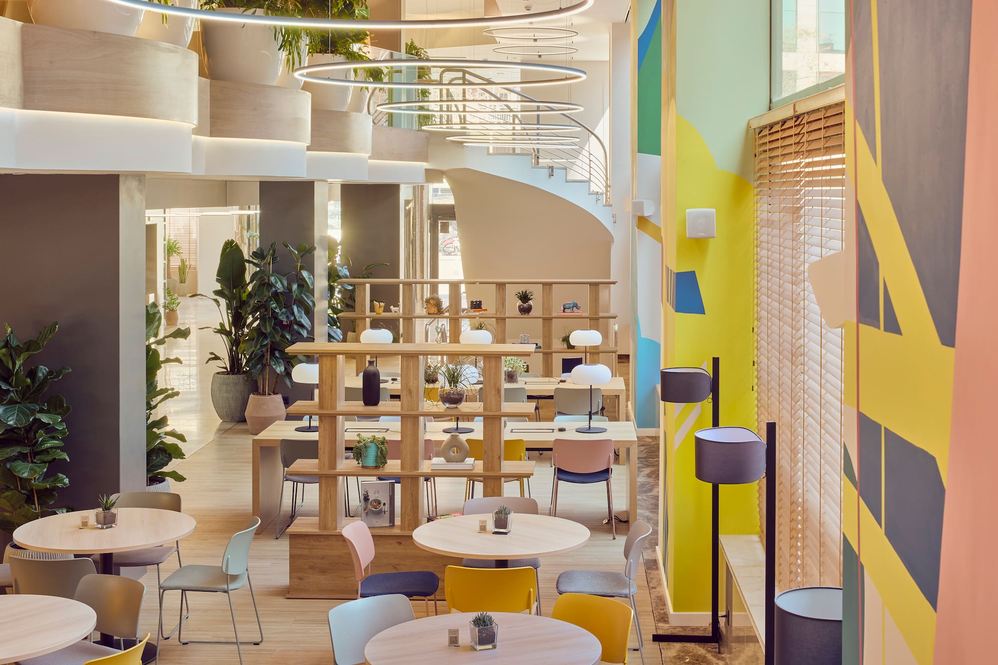 Modern co-working space with colorful walls, wood tables, abundant plants, and a spiral staircase.