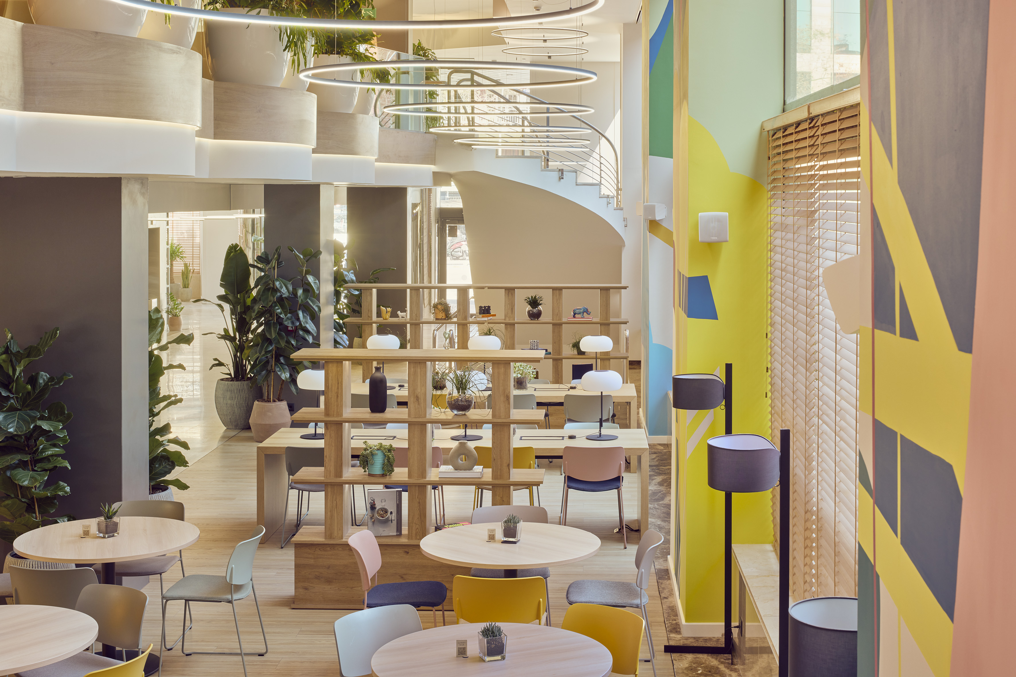 Modern co-working space with colorful walls, wood tables, abundant plants, and a spiral staircase.