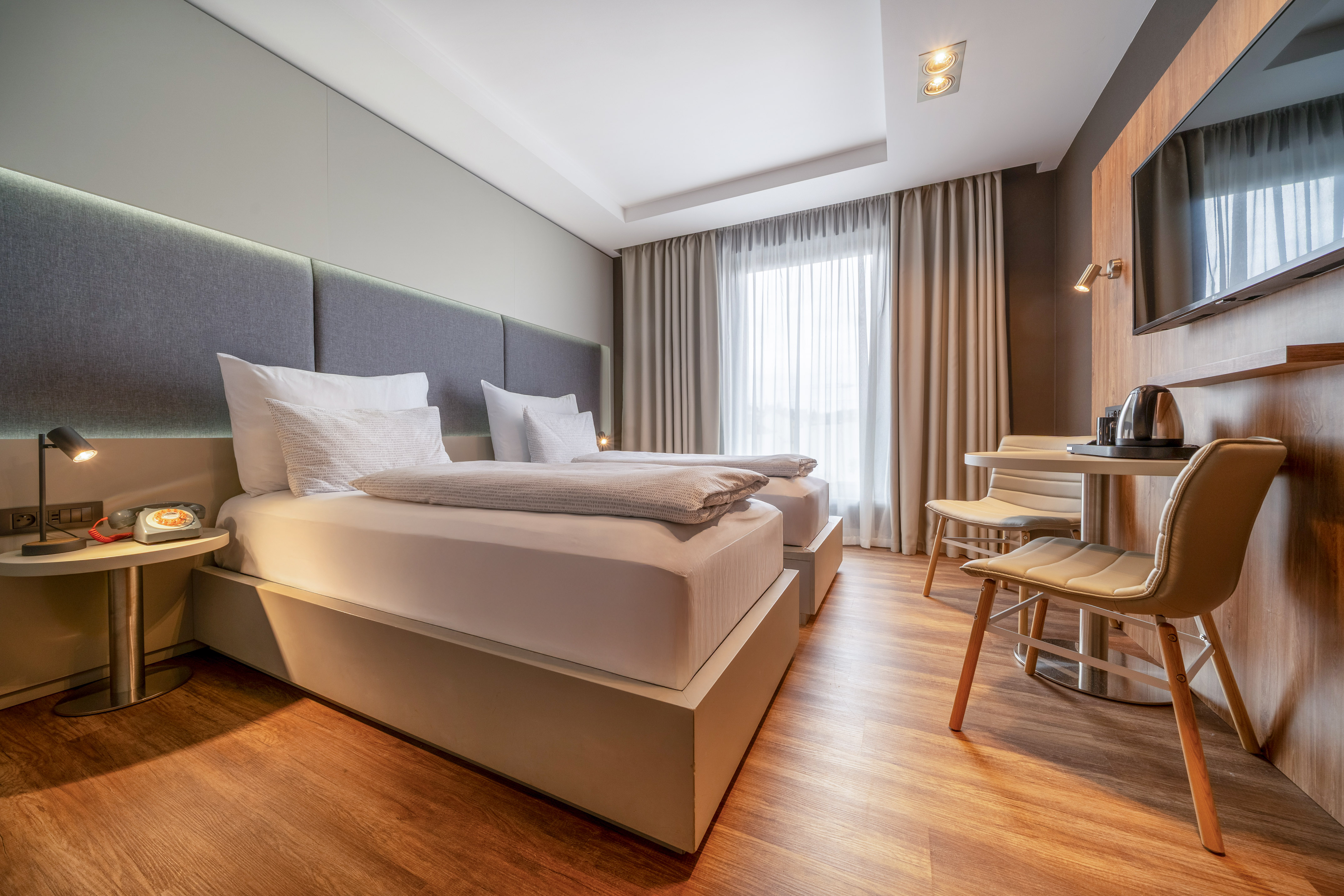 Modern hotel room with twin beds, cozy lighting, wooden floor, TV, and seating.