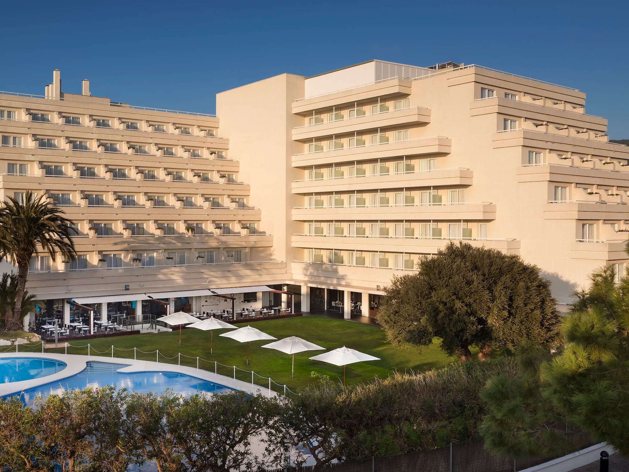 Hotel moderno con piscina y jardín, palmeras y montañas.