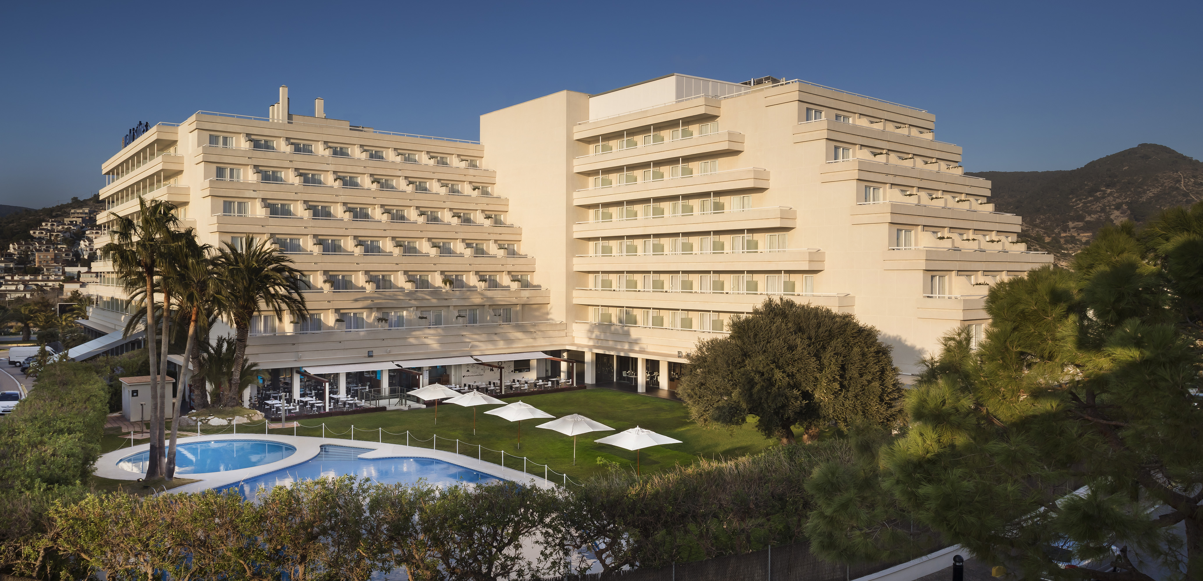 Hotel moderno con piscina y jardín, palmeras y montañas.