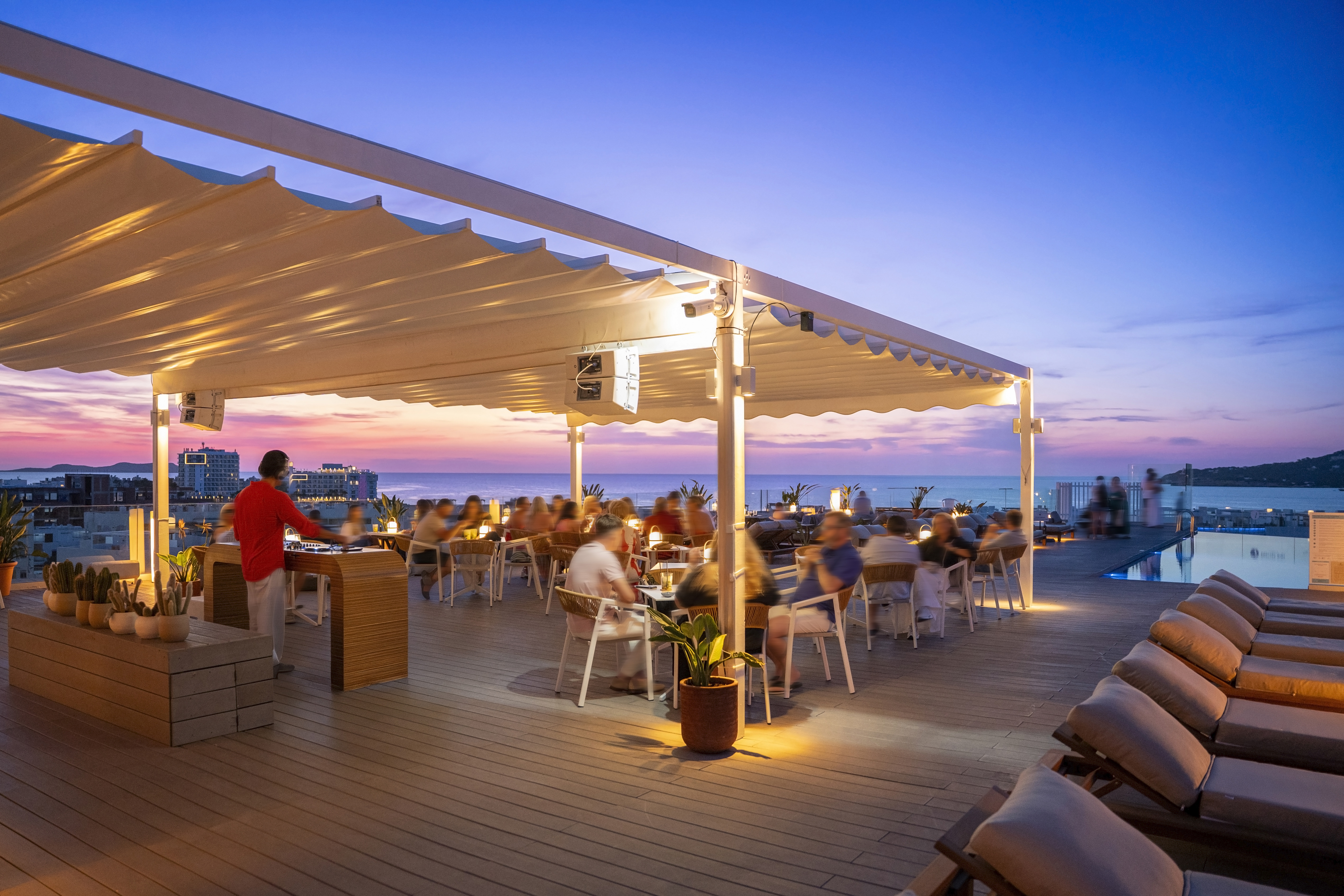 Rooftop bar with dining, DJ, and vibrant sunset ocean view.