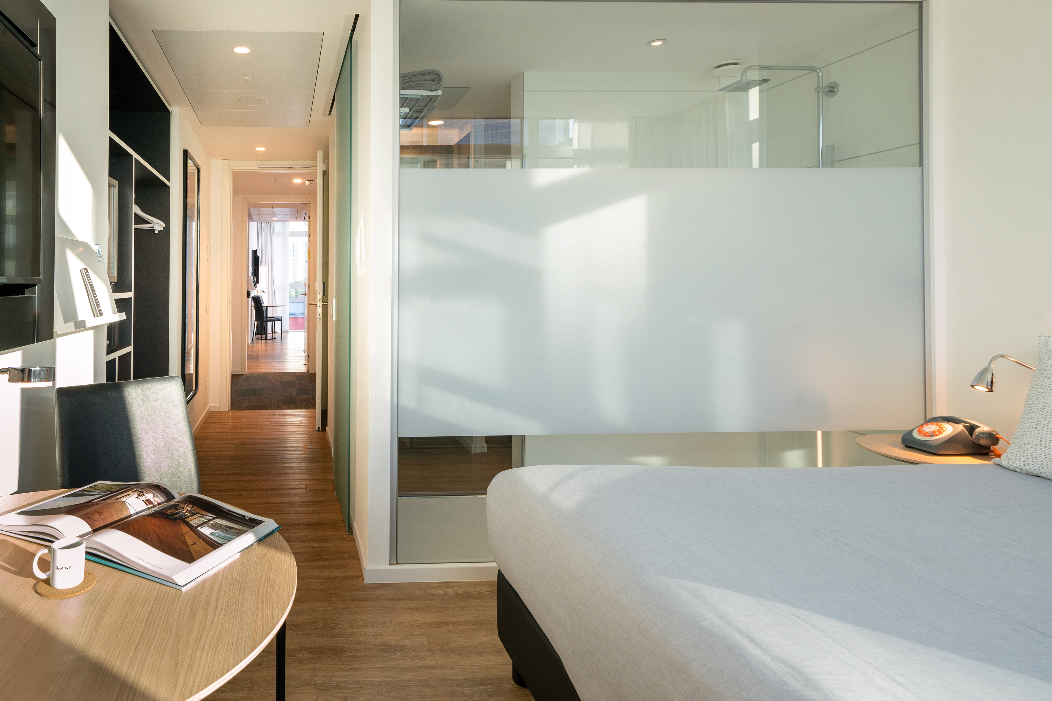 Compact modern hotel room with bed, frosted glass shower, and desk.