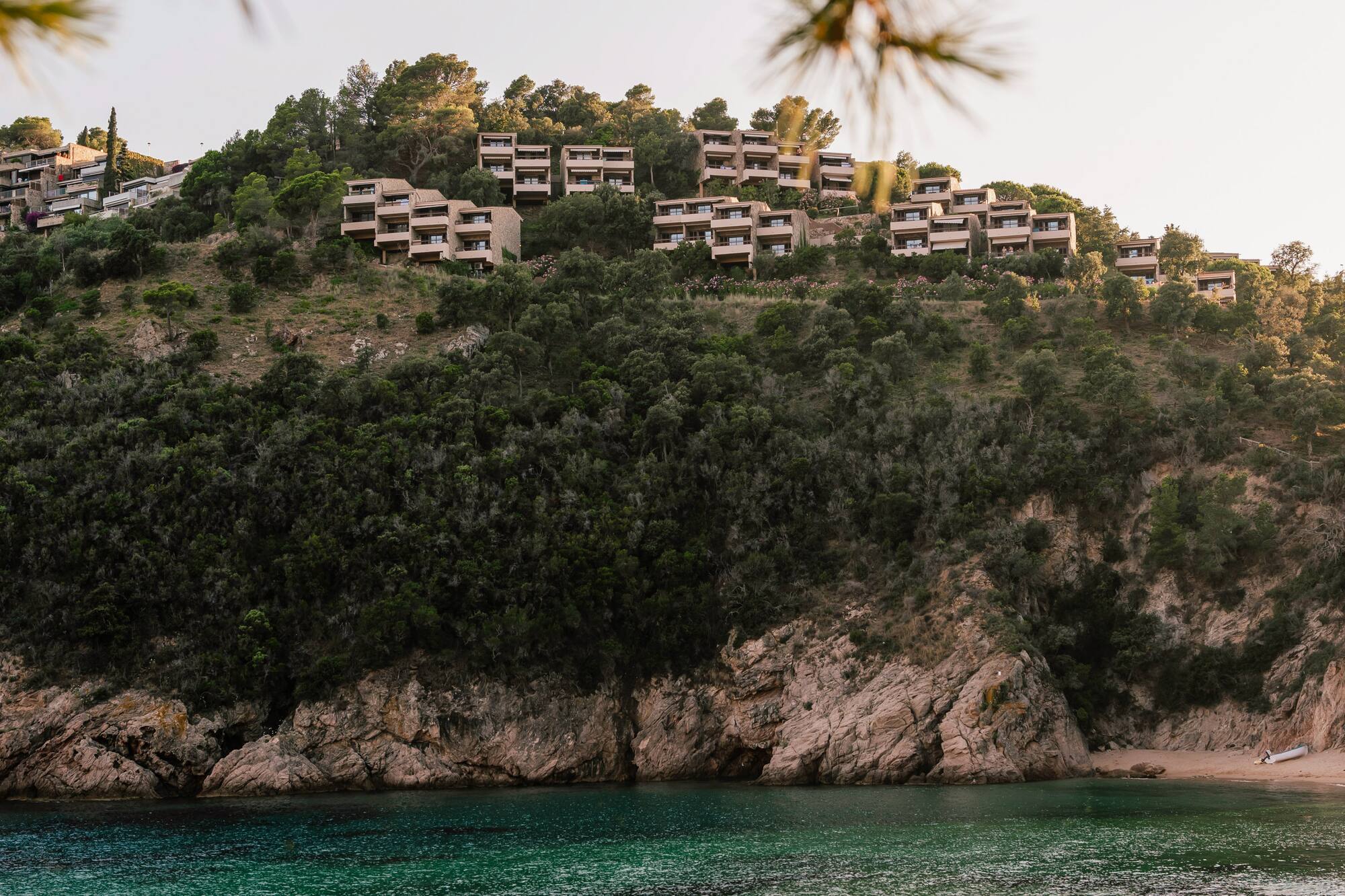 Two paddleboarders in turquoise bay below hillside resort and rocky beach.