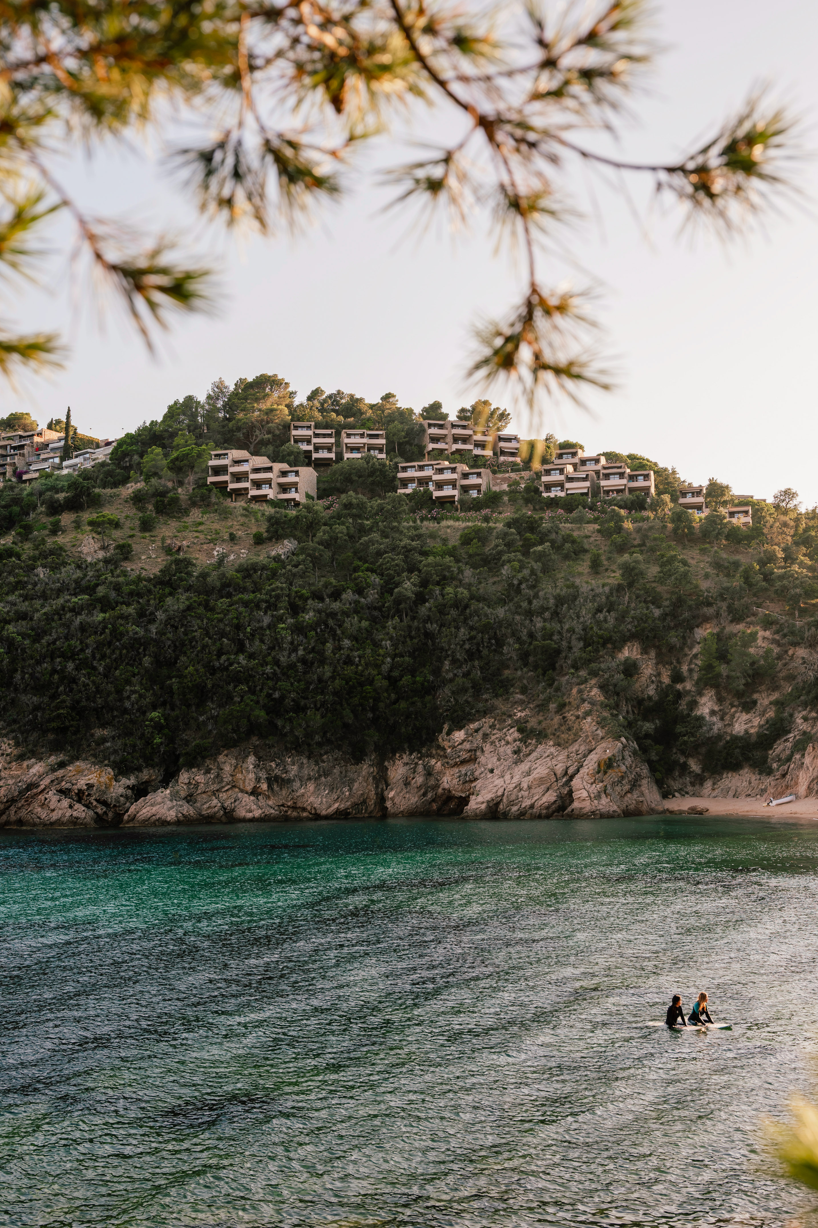 Two paddleboarders in turquoise bay below hillside resort and rocky beach.